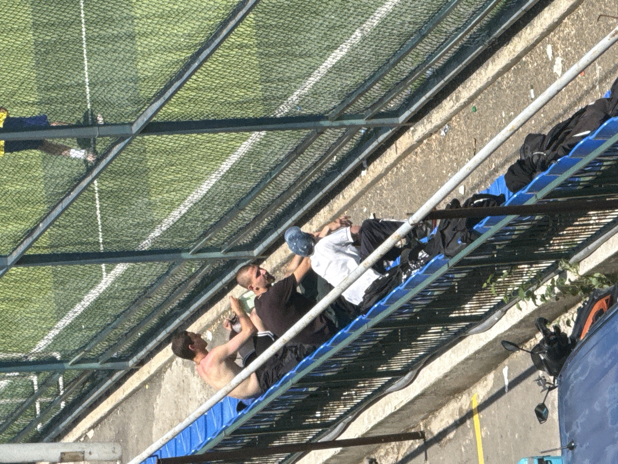 The image depicts a view of several individuals sitting on blue bleachers adjacent to a fenced sports field. In the foreground, the handlebars and mirrors of a parked motorcycle are visible, partially obscuring a blue car. The individuals on the bleachers are dressed casually. One person is shirtless, another wears a dark t-shirt, and a third is in a white t-shirt and a blue cap. Several black bags are placed on the bleachers near the individuals. The sports field appears to be a soccer field with white lines marking its boundaries, and it is partially obscured by a green mesh fence. The overall scene suggests an outdoor urban setting, possibly a sports complex. Based on the provided context, the location is identified as Tbilisi, Georgia.