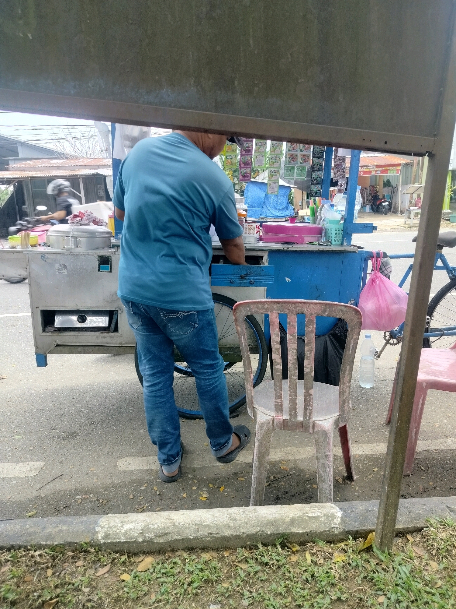 A person stands by a food cart in Kota Tanjung Selor, Indonesia. The person, wearing a blue t-shirt and blue jeans, is facing away from the camera and appears to be preparing food. The food cart is blue and silver, with various containers and equipment visible. A bicycle is attached to the side of the cart. Two chairs, one red and one pink, are positioned in front of the cart. The scene is set on a street with a concrete curb and grassy area. In the background, buildings and a motorcycle are visible, suggesting an urban or semi-urban setting.