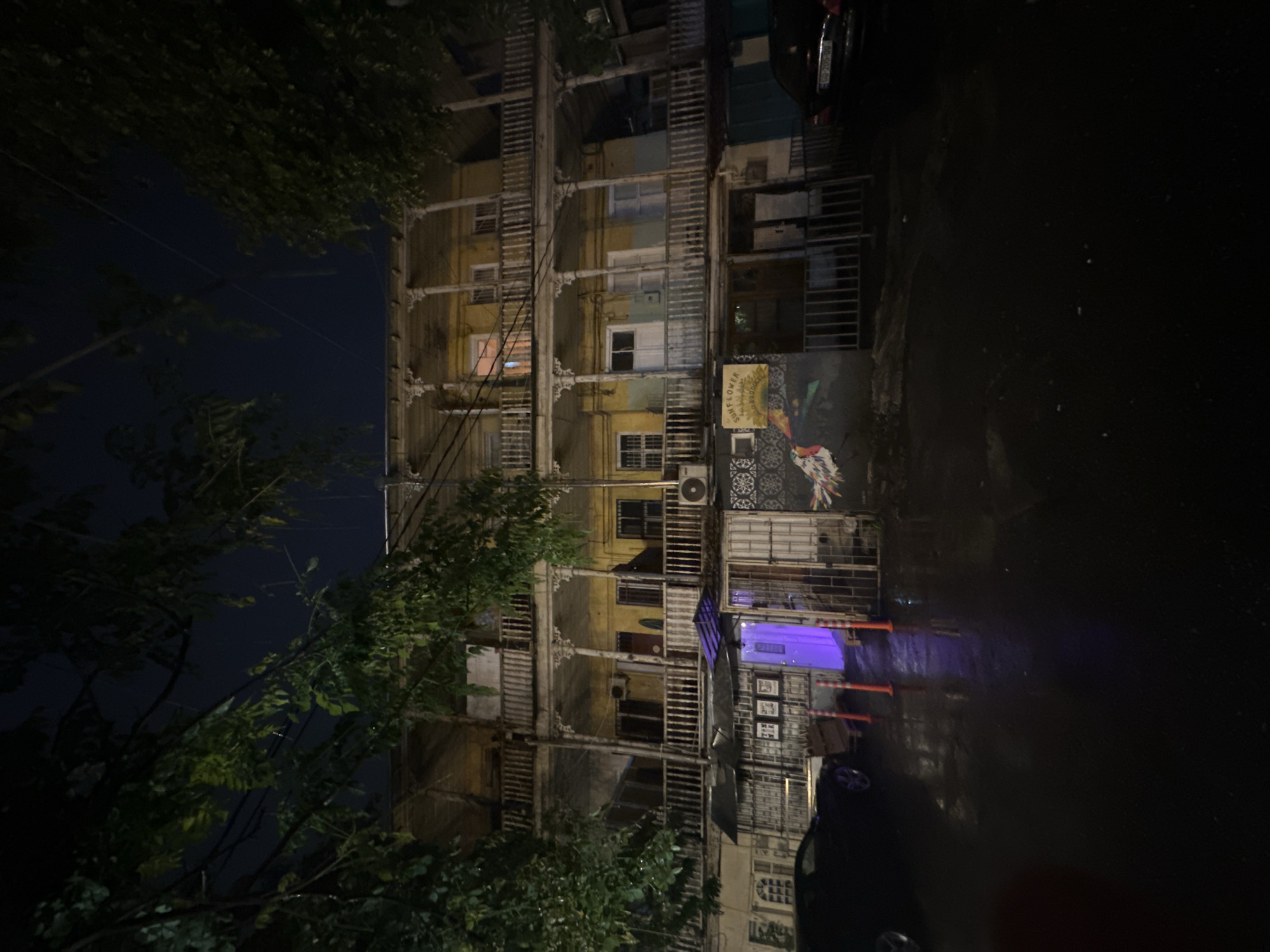 The image depicts a dimly lit urban scene, presumably at night, in an unknown city. The primary subject is a multi-story building with an aged facade, featuring multiple balconies and windows. The building appears to have undergone various additions or renovations over time. A mural or decorative artwork is visible on one side of the building. Street lighting and some artificial lighting within the building provide the only illumination. The street adjacent to the building appears wet, reflecting light, suggesting a recent rain. A car is parked along the street, partially visible. Trees are visible on the left side of the image, obscuring parts of the building and the night sky. Orange traffic cones are lined on the ground. The overall atmosphere is subdued and gritty.