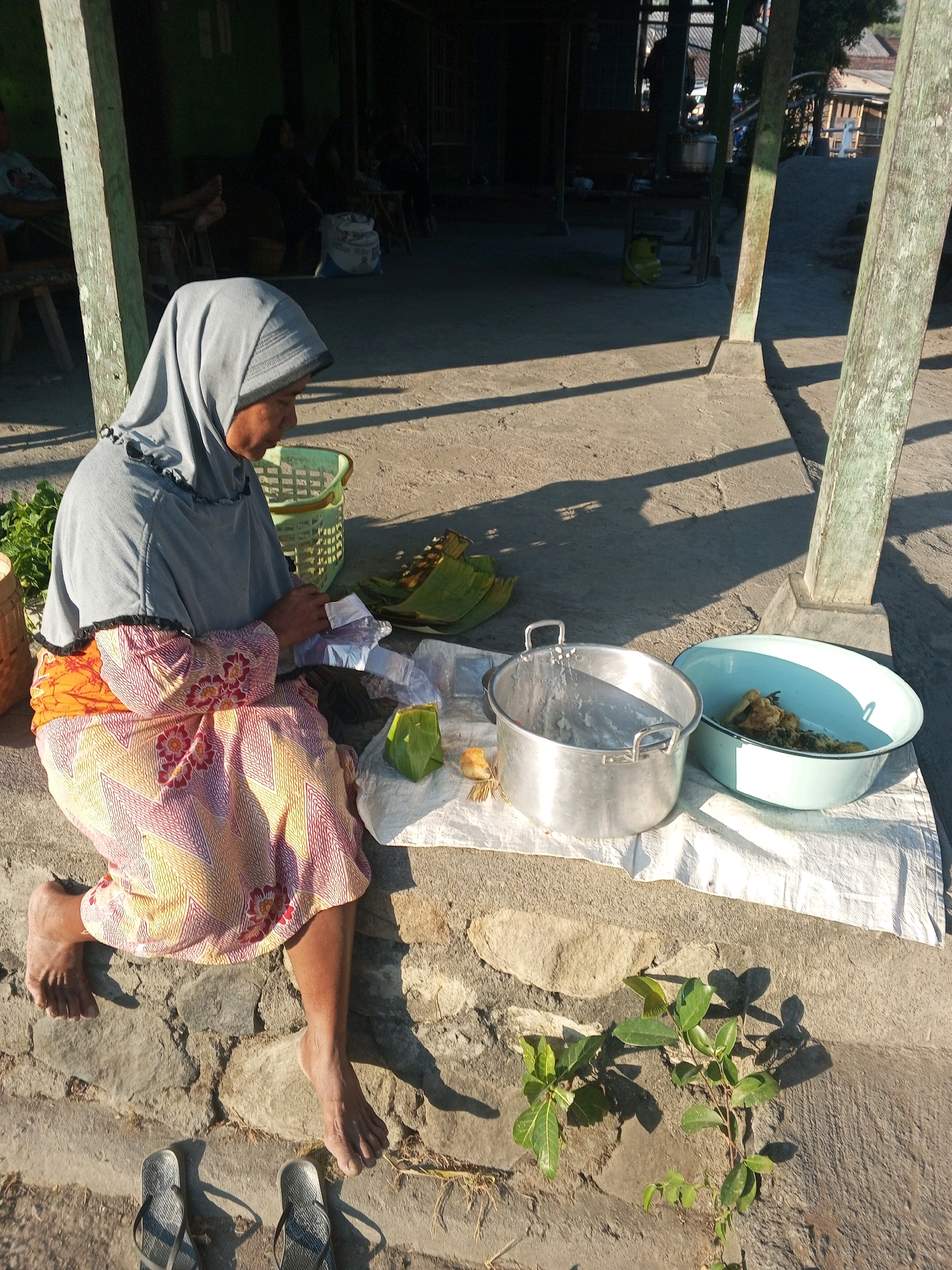 A woman wearing a grey hijab and a colorful patterned dress sits on a stone ledge outdoors. She is looking down at her hands, which are engaged with some small white packets. In front of her are various food items, including some wrapped in banana leaves and a small bird-like object. Two large metal and enamel basins are also present, one empty and the other containing food. Behind her, there is a woven basket and some green plants. The scene appears to be a street or outdoor market setting, possibly in Kedungringin, Indonesia, given the architectural style of the building in the background and the presence of outdoor vendors. The ground is paved and there are shadows cast by the sunlight.
