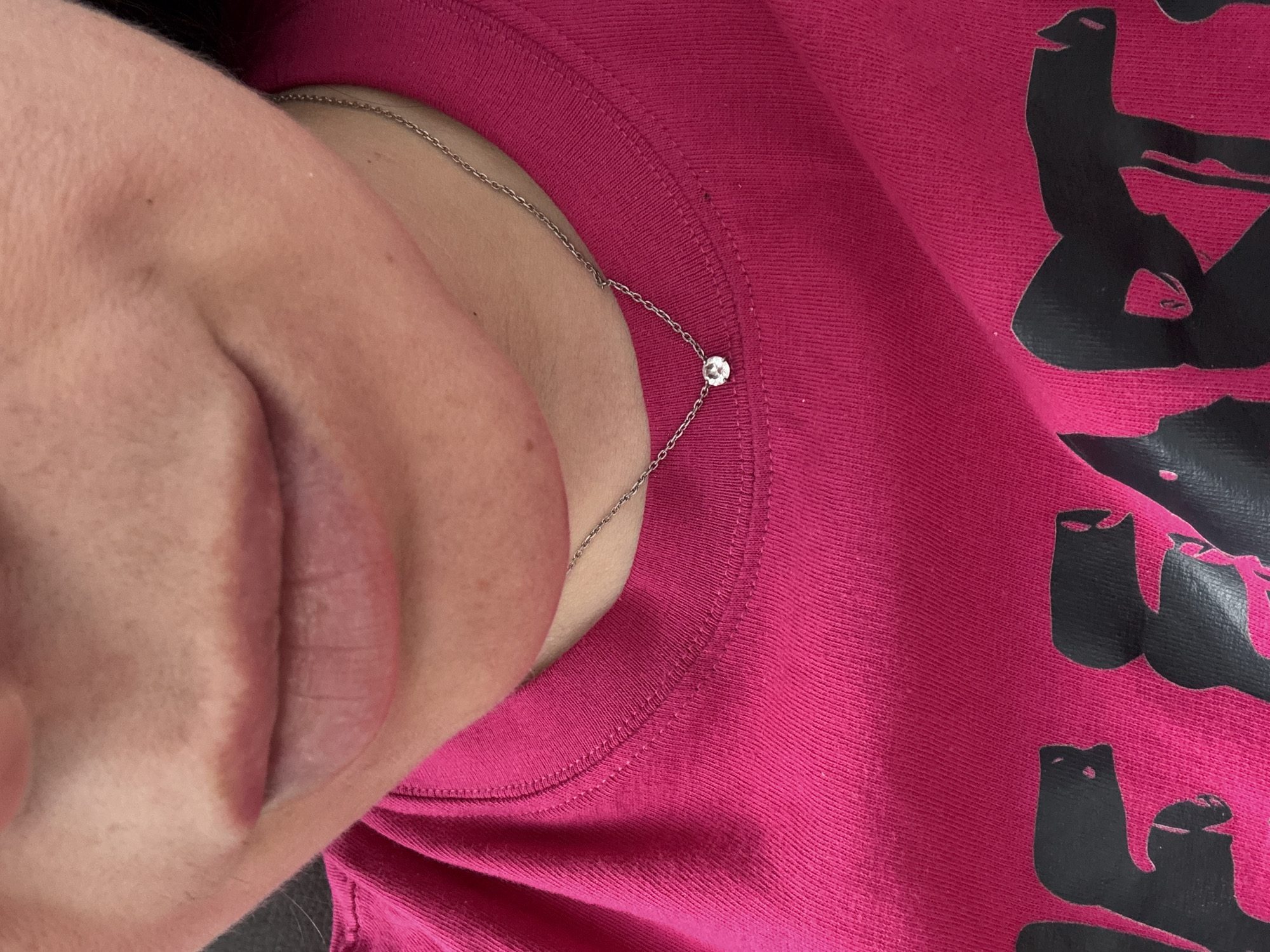 A close-up, tilted shot captures a person's upper body, focusing on their neck, shoulder, and a portion of their face, set against a fuchsia-colored garment. The left side of the frame shows the person's lower face, including their lips and chin, angled towards the top. A delicate, silver-colored chain necklace is visible around their neck, featuring a single, small, round, clear gemstone, likely a diamond or cubic zirconia, set into the chain near the collarbone.
The fuchsia garment, appearing to be a t-shirt or sweatshirt, exhibits a textured knit fabric and visible stitching along the neckline and shoulder area. On the right side of the image, a partially visible black graphic design or stylized text is printed on the fuchsia fabric. The shapes are abstract, some resembling parts of letters (like 'E' or 'L') or simplified figures that could be interpreted as cat-like shapes.
The photograph is a portrait-style shot, highlighting the subject's jewelry and clothing. No specific activity or event is depicted beyond posing for the image. The setting is indistinct due to the close crop. No time of day or weather conditions can be determined from the indoor context. The visible facial features do not convey strong emotions. The context provided for this image is Chaikovsky, Russia.