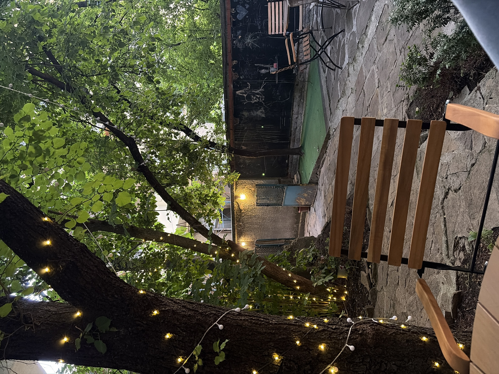 The image depicts an outdoor patio or garden area at dusk, adorned with string lights. The ground is paved with irregular stone tiles. In the foreground, a wooden chair with a slatted back and armrest is visible. Behind it, another wooden chair is partially obscured. To the left, the trunk of a large tree is wrapped with string lights, illuminating its bark and the surrounding foliage. Further into the scene, a black wall is covered with chalk drawings. A small patch of artificial green turf is situated in front of the wall. Several metal chairs and tables are arranged on the patio. The overall atmosphere suggests a casual, possibly restaurant or cafe setting, illuminated by the warm glow of the string lights.