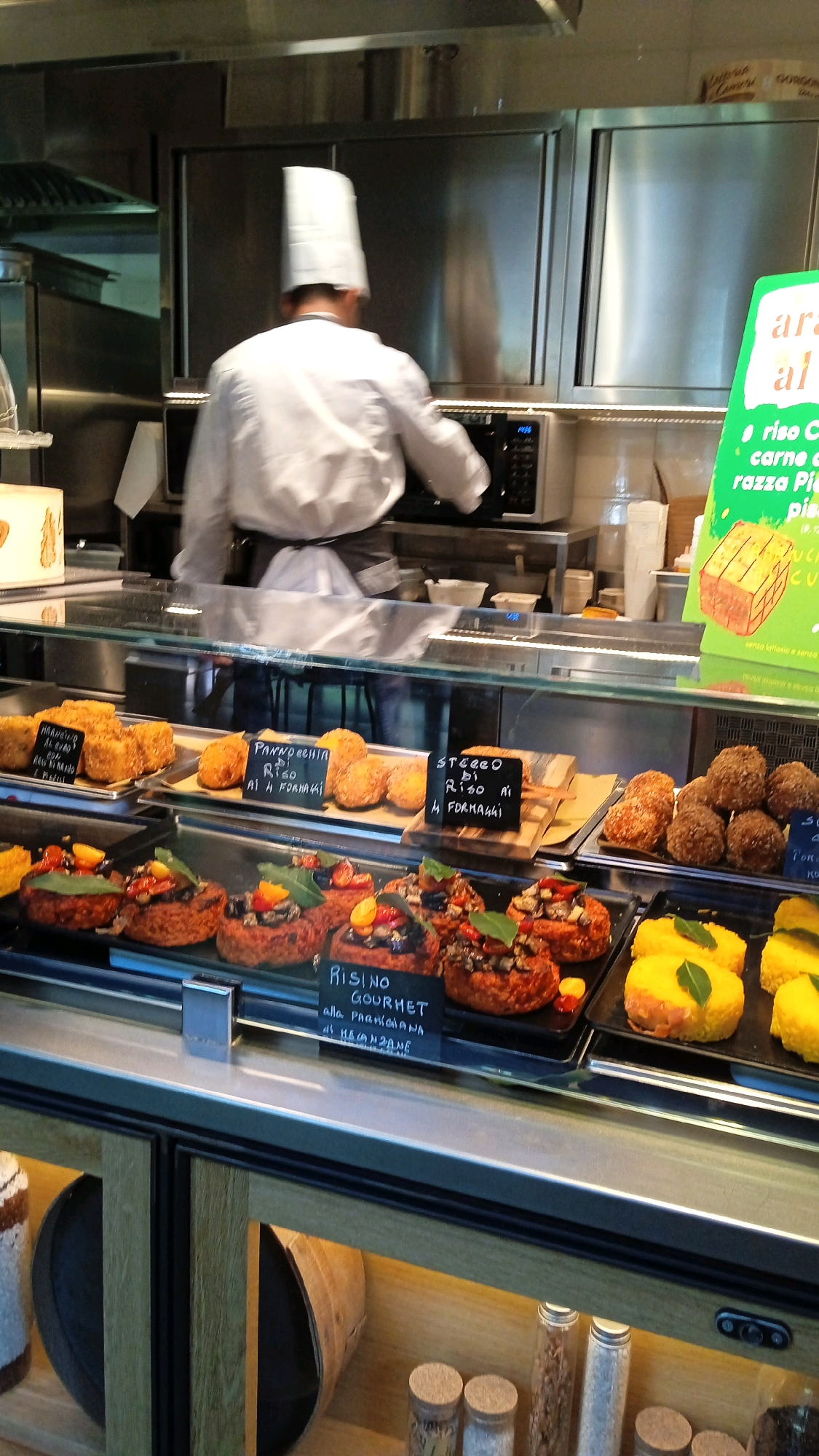 A chef in a white uniform and tall hat is seen from the back, operating a microwave oven in a commercial kitchen. The kitchen features stainless steel surfaces and equipment. In the foreground, a display case holds various food items. These include several golden-brown fried balls and what appear to be small, savory cakes topped with vegetables and herbs, arranged on black trays. To the right, bright yellow, rectangular food items are also on display, garnished with green leaves. A price tag in Italian reads "RISOTTO GOURMET alla Parmigiana di Raviolì". Another sign in green, also in Italian, advertises "arance" and mentions "riso carne di razza Piedmontese", with an image of a block of cheese. The overall scene suggests a food preparation or serving area within a restaurant or deli in Milan, Italy.