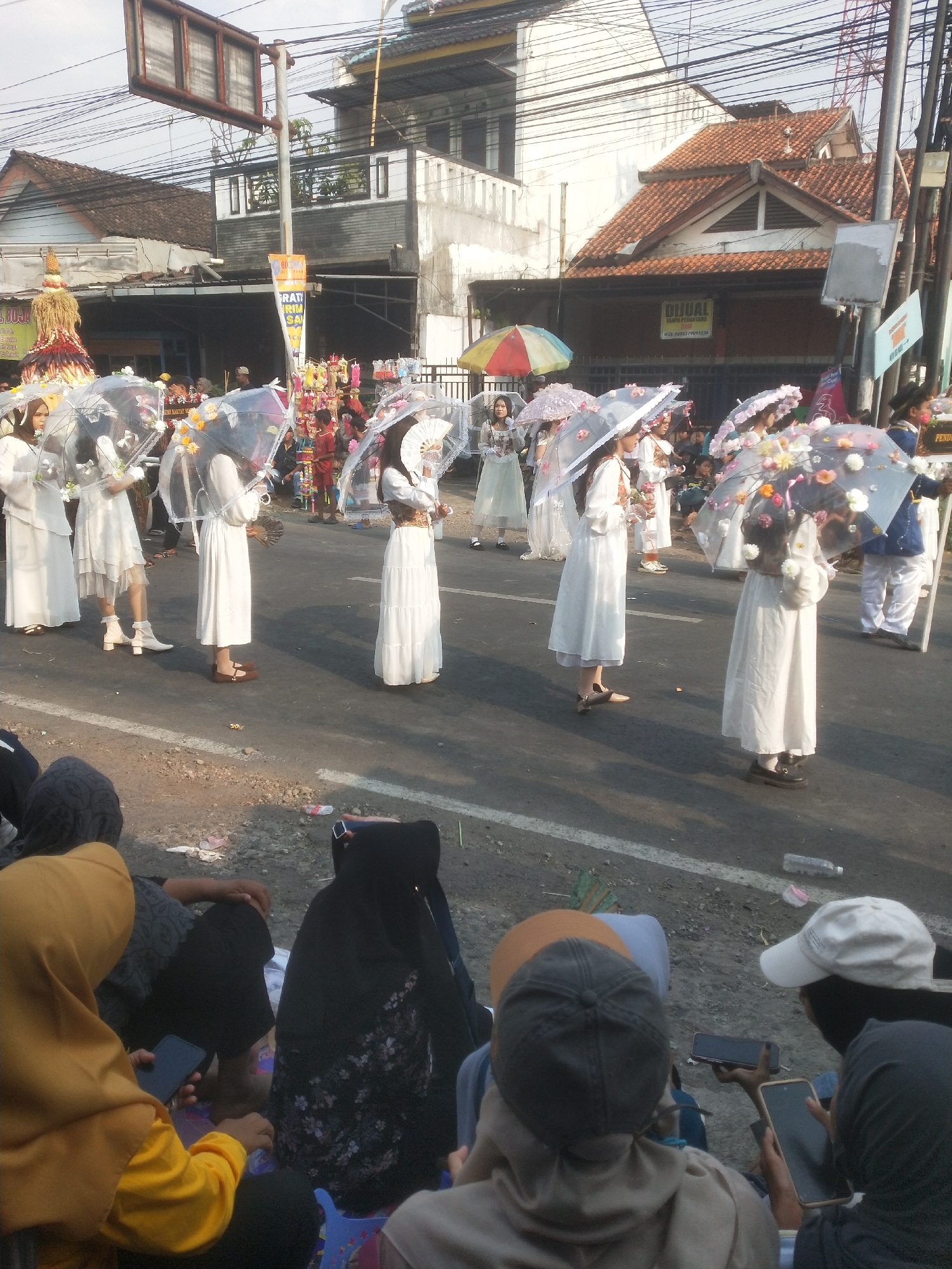 In Boja, Indonesia, a procession is underway, featuring several young women dressed in white dresses, some holding ornate umbrellas. The women are walking on a paved road, flanked by a crowd of onlookers sitting on the roadside. The buildings in the background appear to be a mix of residential and commercial structures, with a visible power line crossing overhead. The atmosphere suggests a local festival or cultural event.