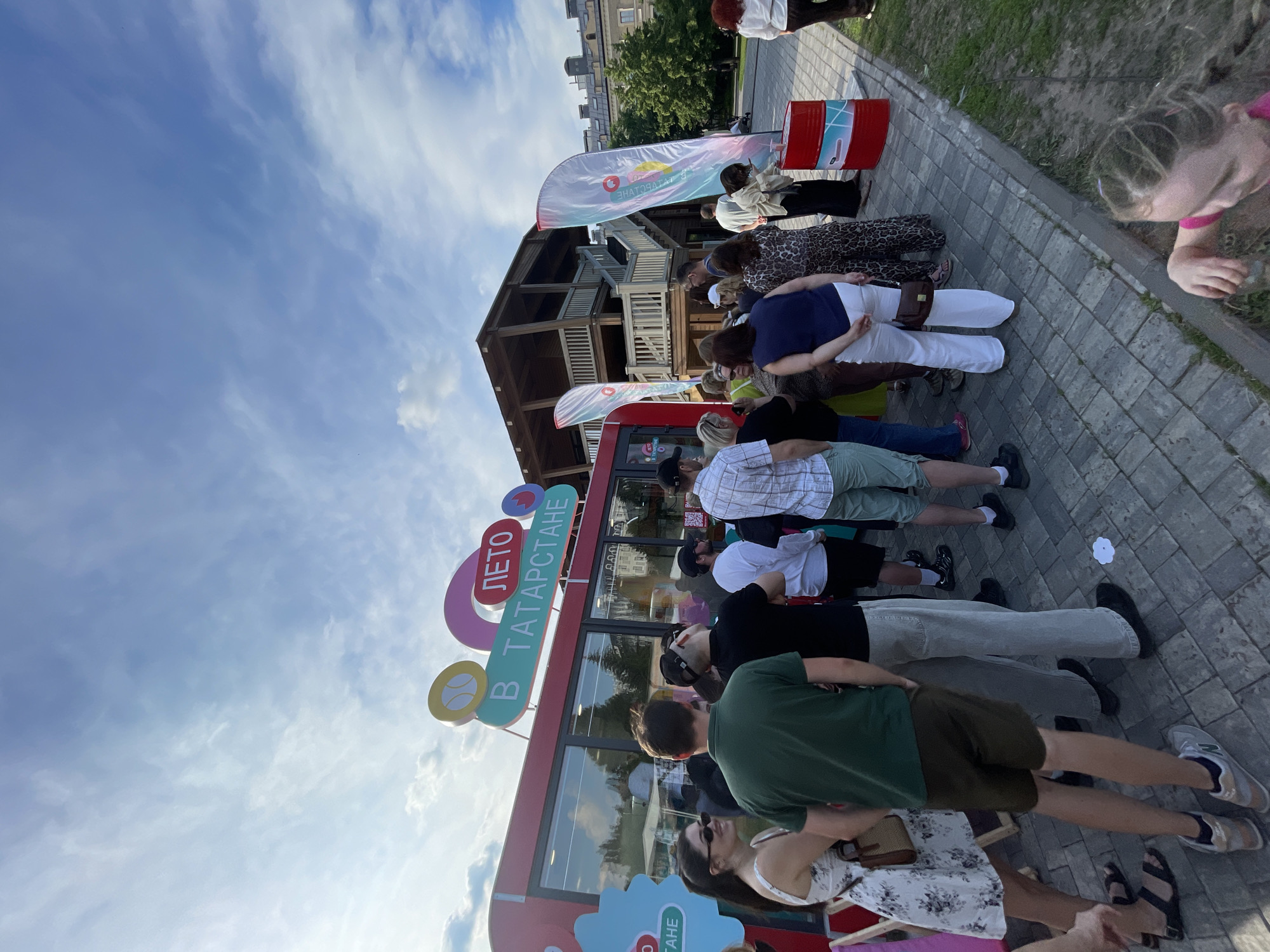 The image depicts a lively outdoor scene in Kazan, Russia, possibly near the Kazan Kremlin, judging by the architectural style of the building in the background. In the foreground, a diverse group of people are gathered, many of them standing in a queue. A bright red kiosk with a sign that reads "ЛЕТО В ТАТАРСТАНЕ" (Summer in Tatarstan) is visible, suggesting a promotional event or a summer-themed attraction. Several people are casually dressed, indicating a warm day. Among the objects present are two tall, white flag banners with colorful logos, a red oil drum with a white and blue design, and a woman in a white dress in the foreground. The ground is paved with grey interlocking bricks, and there is a grassy area to the right. The sky is a clear blue with scattered white clouds. The overall atmosphere appears to be one of a public gathering or festival.