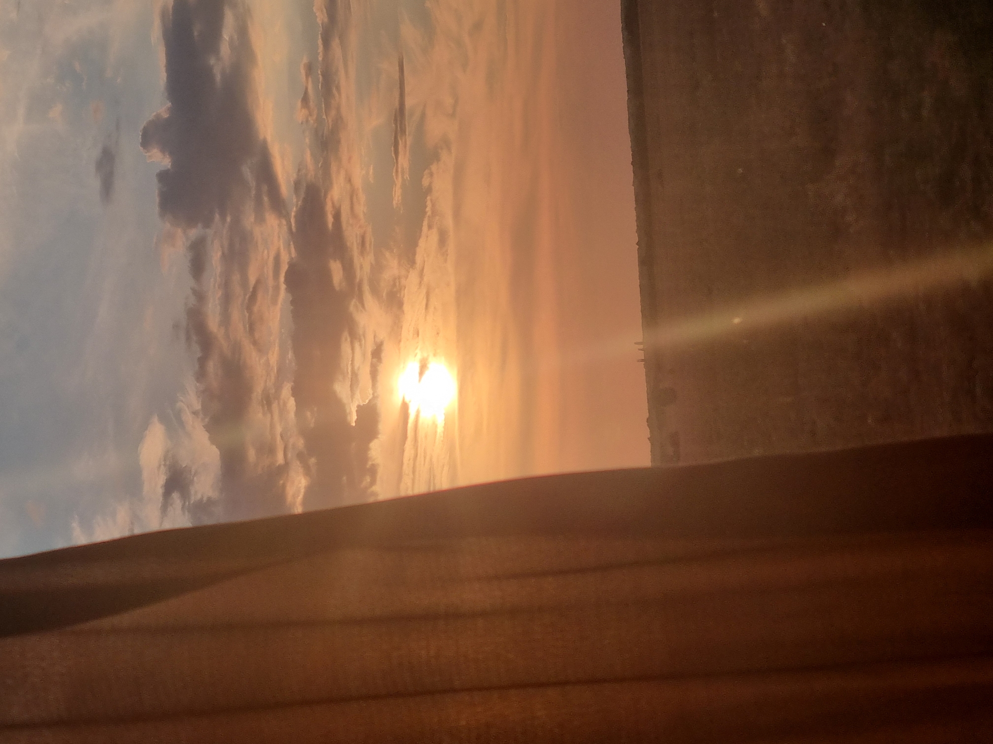 The image depicts a sunset viewed through a window with a curtain partially obscuring the view. The sky is filled with warm hues of orange, pink, and purple, with scattered clouds catching the light. The sun is visible as a bright, glowing orb near the center of the frame. Below the sky, a landscape stretches out, appearing to be fields or plains with a gentle rise in the distance. There are a few faint, dark vertical shapes on the horizon, possibly trees or structures. The foreground is dominated by a soft, textured curtain, likely made of fabric, with visible folds and a warm, earthy tone. The overall impression is one of a serene and natural scene, possibly captured from a residential or hotel window. The question asks to identify the location in Avram Iancu, Romania, however, the image itself does not provide any specific geographical markers or context to confirm this location.