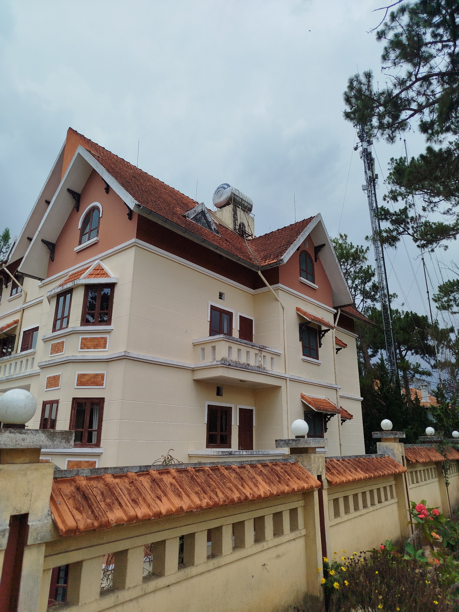 A residential building with a beige facade and red tiled roof is seen from a low angle under an overcast sky in Da Lat, Vietnam. The building is two stories tall with an attic level. It has multiple windows with dark brown frames and some windows have small awnings. A balcony is visible on the second floor. The building is surrounded by a low wall topped with matching red tiles and decorative posts with spherical lights on top. Trees and a tall telecommunications tower are visible in the background. Flowers and vegetation grow in front of the wall.