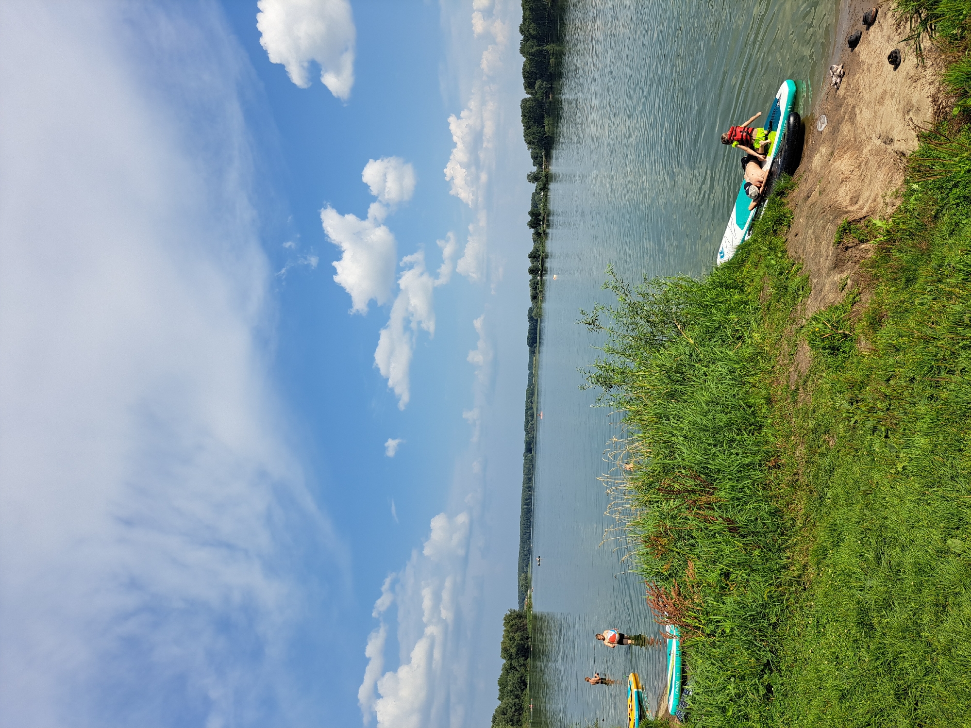 The image depicts a wide, calm lake with a tree-lined shore in the distance under a bright, cloudy sky. In the foreground, lush green grass and vegetation lead down to a sandy bank where two people are on a paddleboard in the shallow water. One person, wearing a life vest, is kneeling on the paddleboard, while another person, also wearing a life vest, is lying down. Several other people are visible in the water and on paddleboards further out in the lake. The scene suggests a recreational day at a lake, possibly a park or designated swimming area, in or near Sorochii Log, Russia.