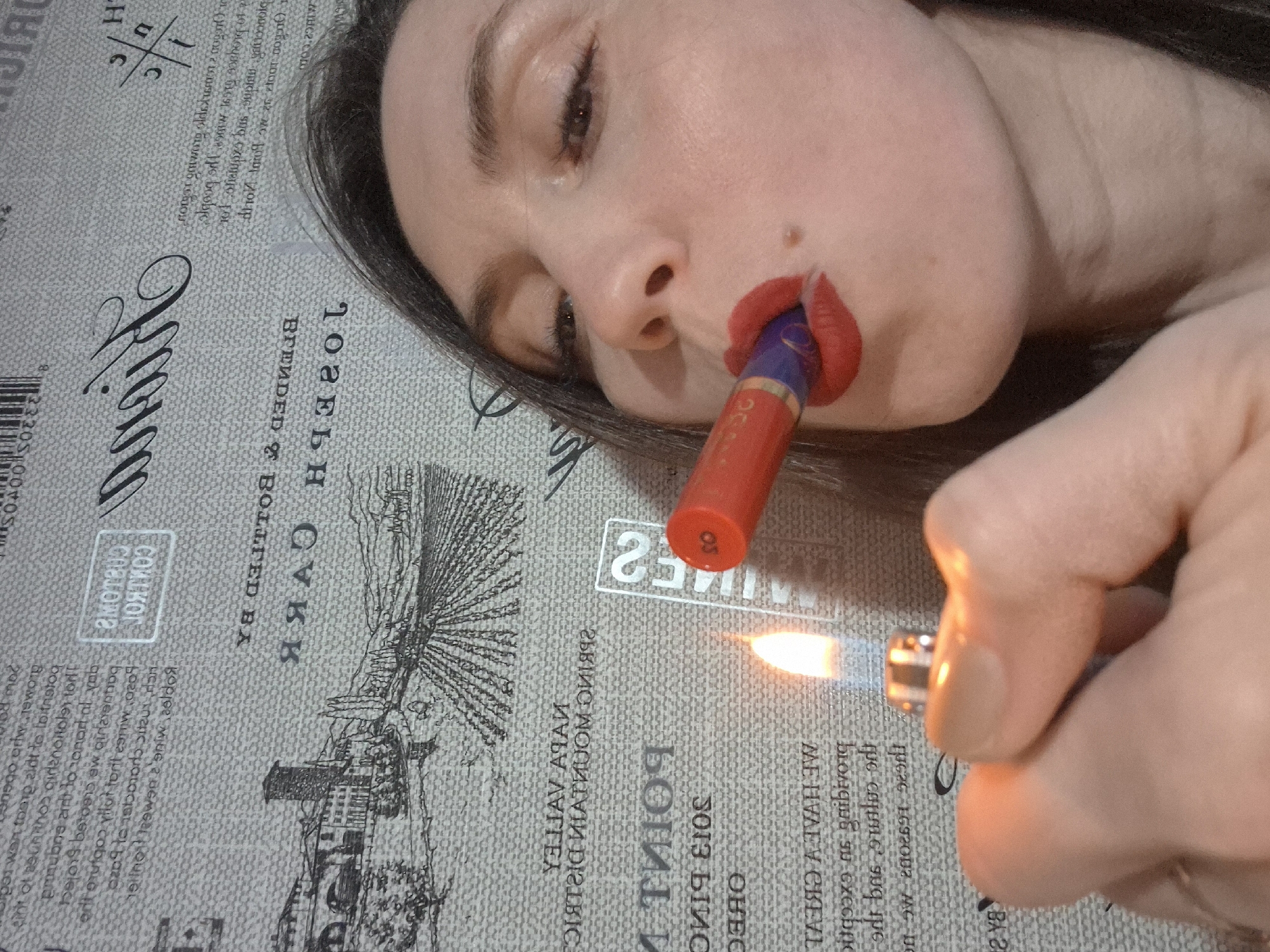 The image depicts a person, presumably female, lying on a printed surface. The person is holding a lit lighter near a tube of lipstick, which is held in their mouth. The printing on the surface appears to be text and illustrations, possibly related to wine or spirits. The lighter is held in the right hand, and the flame is directed towards the end of the lipstick tube. The person's face is partially visible, showing red lips, and the overall scene appears to be indoors.