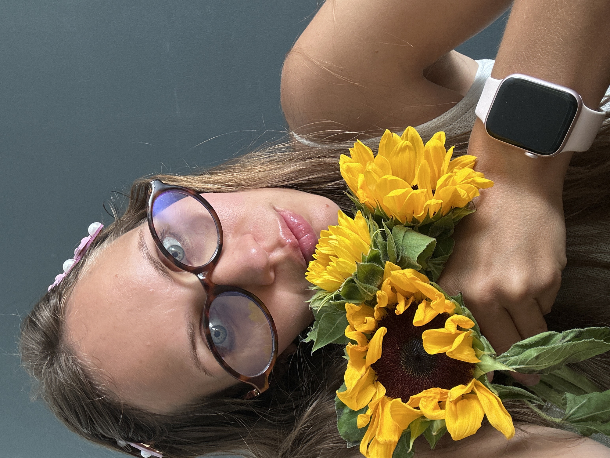 A young woman with brown hair, wearing glasses and a pink headband, is holding a bouquet of sunflowers. She is also wearing a smartwatch on her wrist. The background appears to be a solid gray wall. There is no indication of the location being Fethiye, Turkey.