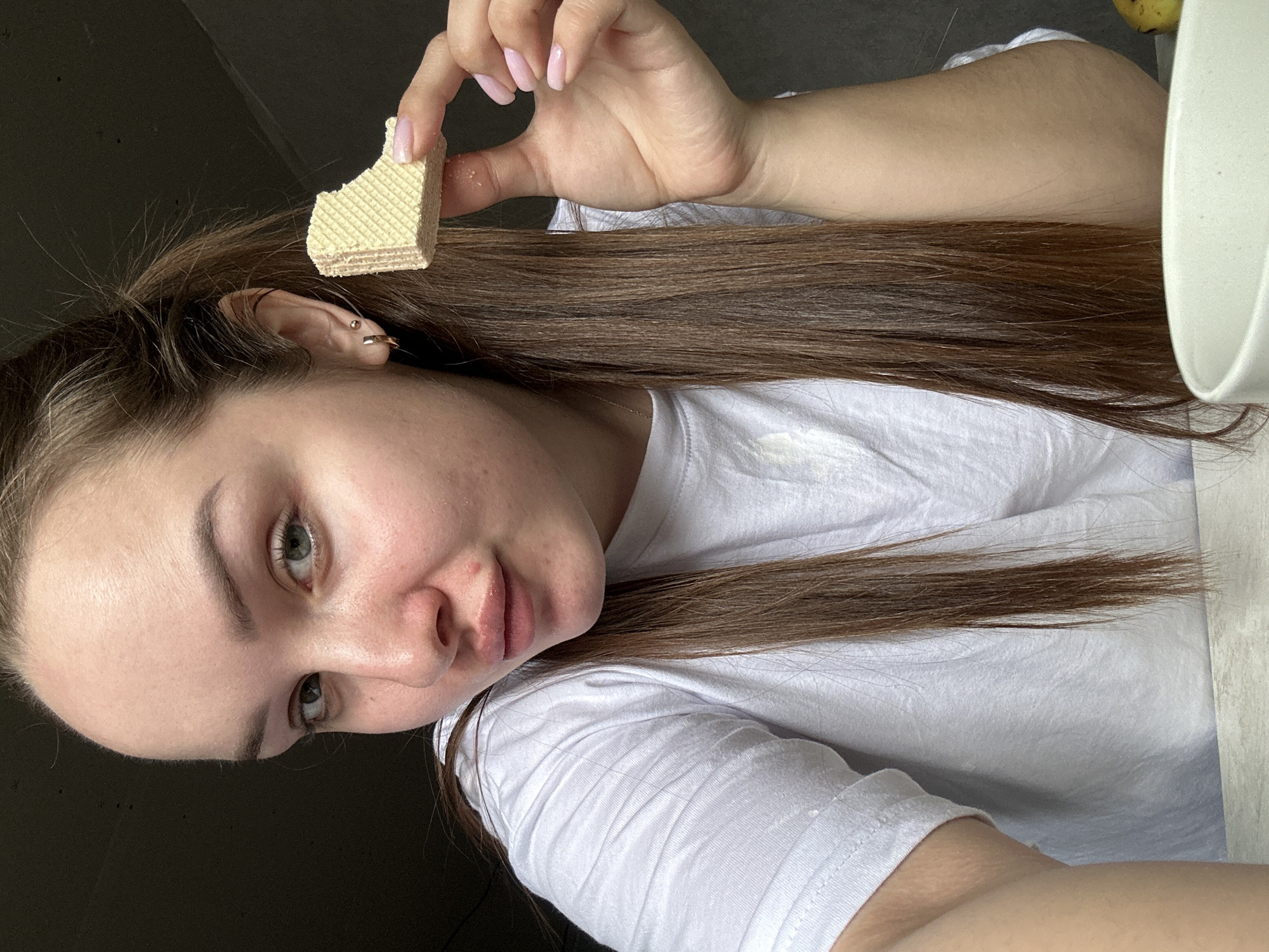 A woman with light skin, long brown hair, and green eyes is holding a piece of a square wafer cookie in her right hand. She is wearing a white t-shirt. Her head is tilted towards the camera. She has piercings in her left ear. The background is dark, with part of a white mug and a yellow object visible at the top right. There is no indication of the location being Ludorvay, Russia.