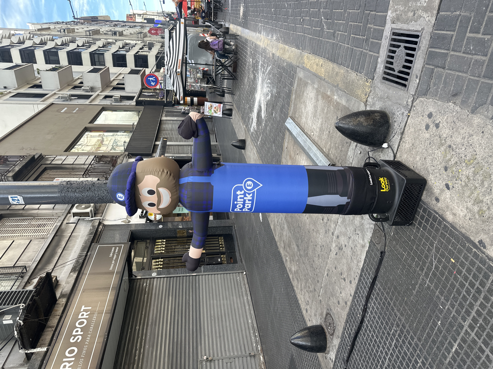 A street scene in Buenos Aires, Argentina, includes an inflated advertising figure that is shaped like a cartoon construction worker in blue with the words "Point Park" printed on it. The advertisement is standing on a gray, tiled sidewalk with a black metal grate for drainage, anchored by a black fan base connected to an electrical cord. The storefronts behind the advertisement have closed metal shutters, and a sign above the storefront reads "RIO SPORT." Across the street are buildings with multiple floors and modern architectural design, and a restaurant awning with a black and white striped pattern. Two people are sitting at an outside table. A no parking sign in Spanish is visible near the restaurant.