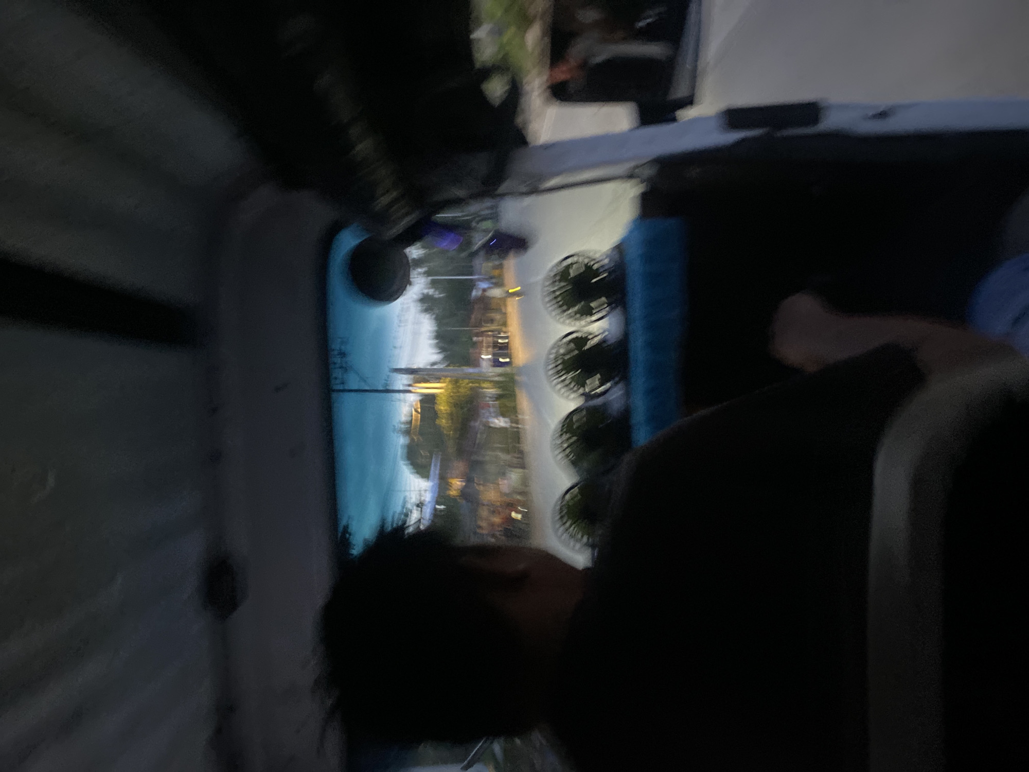 An over-the-shoulder shot from inside a vehicle shows the head and shoulders of a person with dark hair, facing forward towards the windshield. The person is wearing a black t-shirt. Mounted on the dashboard in front of them are four small, round electric fans. The windshield offers a view of a street scene at dusk or dawn. Buildings and trees are visible, with some lights illuminated. The image appears to be taken in Davao City, Philippines, indicated by the presence of similar vehicles and the surrounding environment typical of the city.