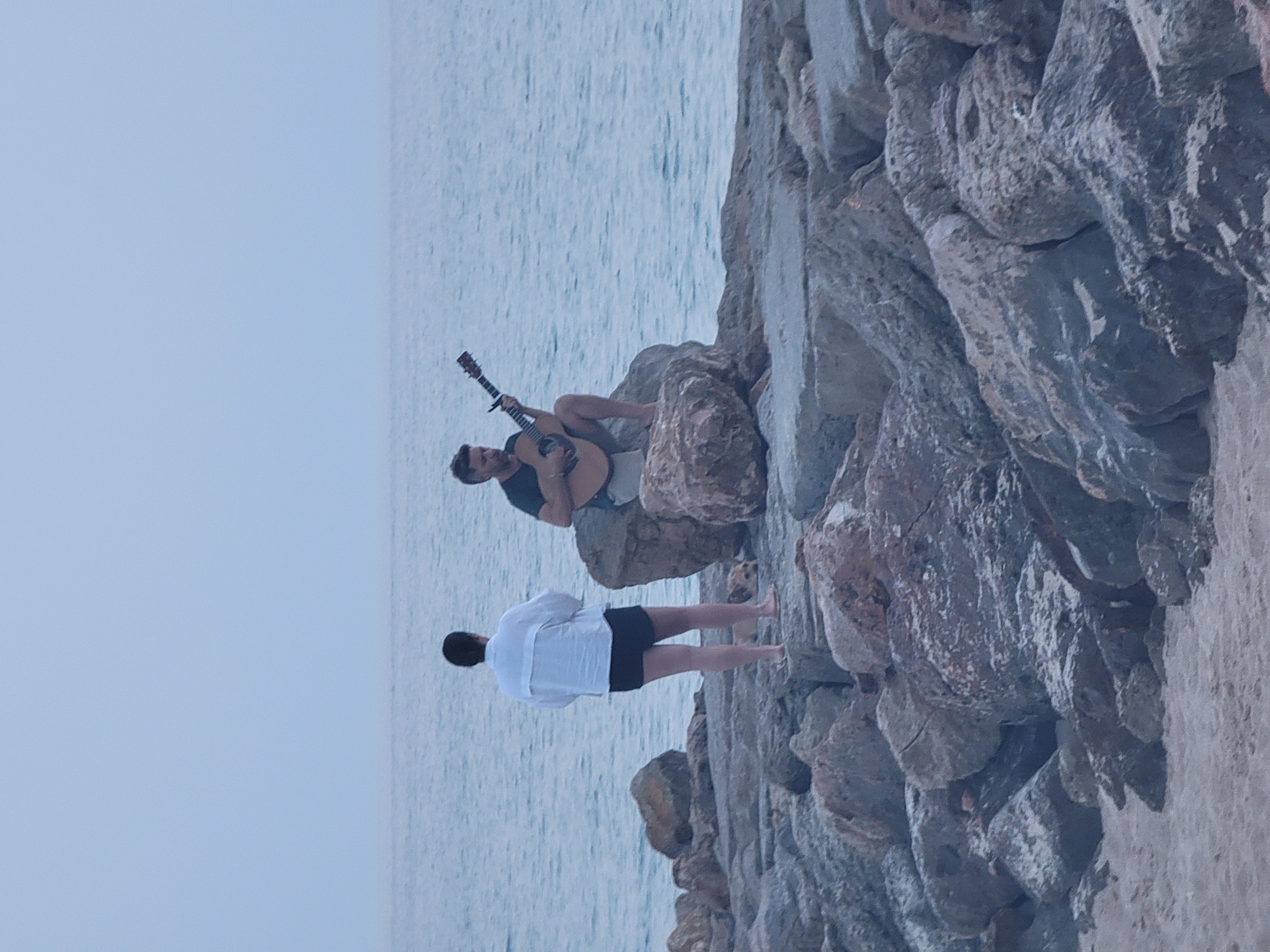 A man sits on a rock by the ocean in Quarteira, Portugal, playing an acoustic guitar. A woman stands facing the ocean, with her back to the camera, and a small dog is lying on the rocks between them. The scene takes place on a rocky shoreline with the ocean stretching out to the horizon under an overcast sky.