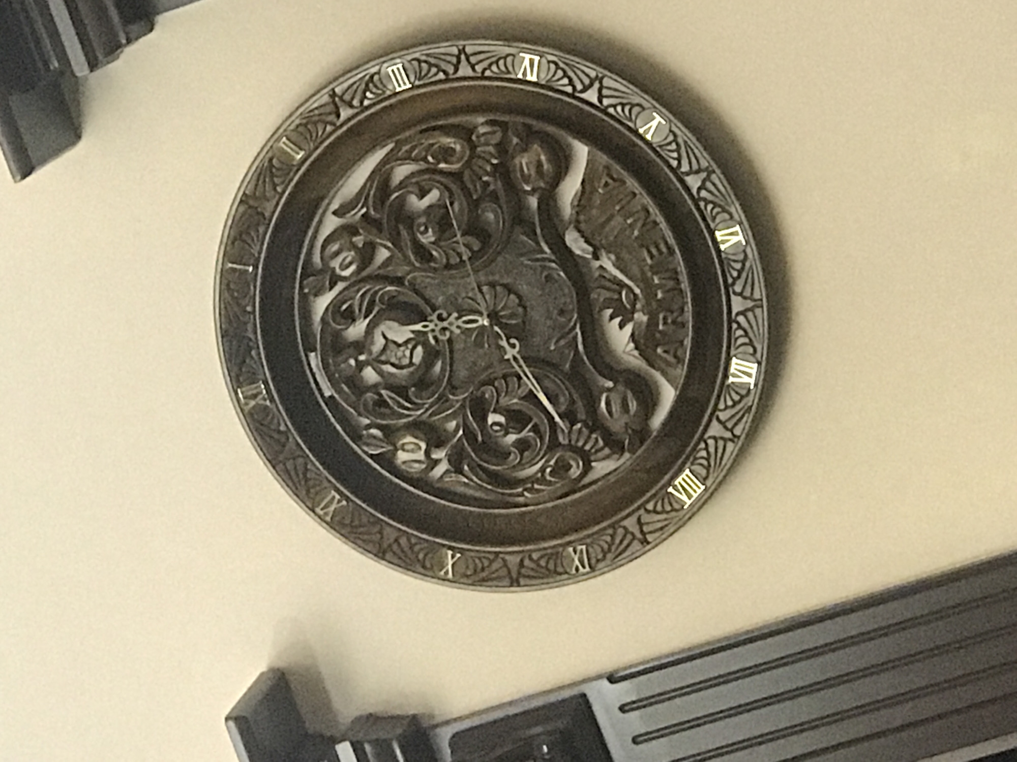 The image depicts a clock hanging on a wall. The clock face is ornate, featuring Roman numerals marking the hours and the word "ARMENIA" inscribed in the clock's center. The clock appears to be made of metal, possibly bronze or a similar alloy. The time displayed is approximately 4:51. The presence of "ARMENIA" on the clock suggests it may be located in Yerevan, Armenia, or at least be associated with Armenian culture. The clock hangs on a light-colored wall. Additional objects, possibly parts of furniture or fixtures, are visible in the corners of the frame.