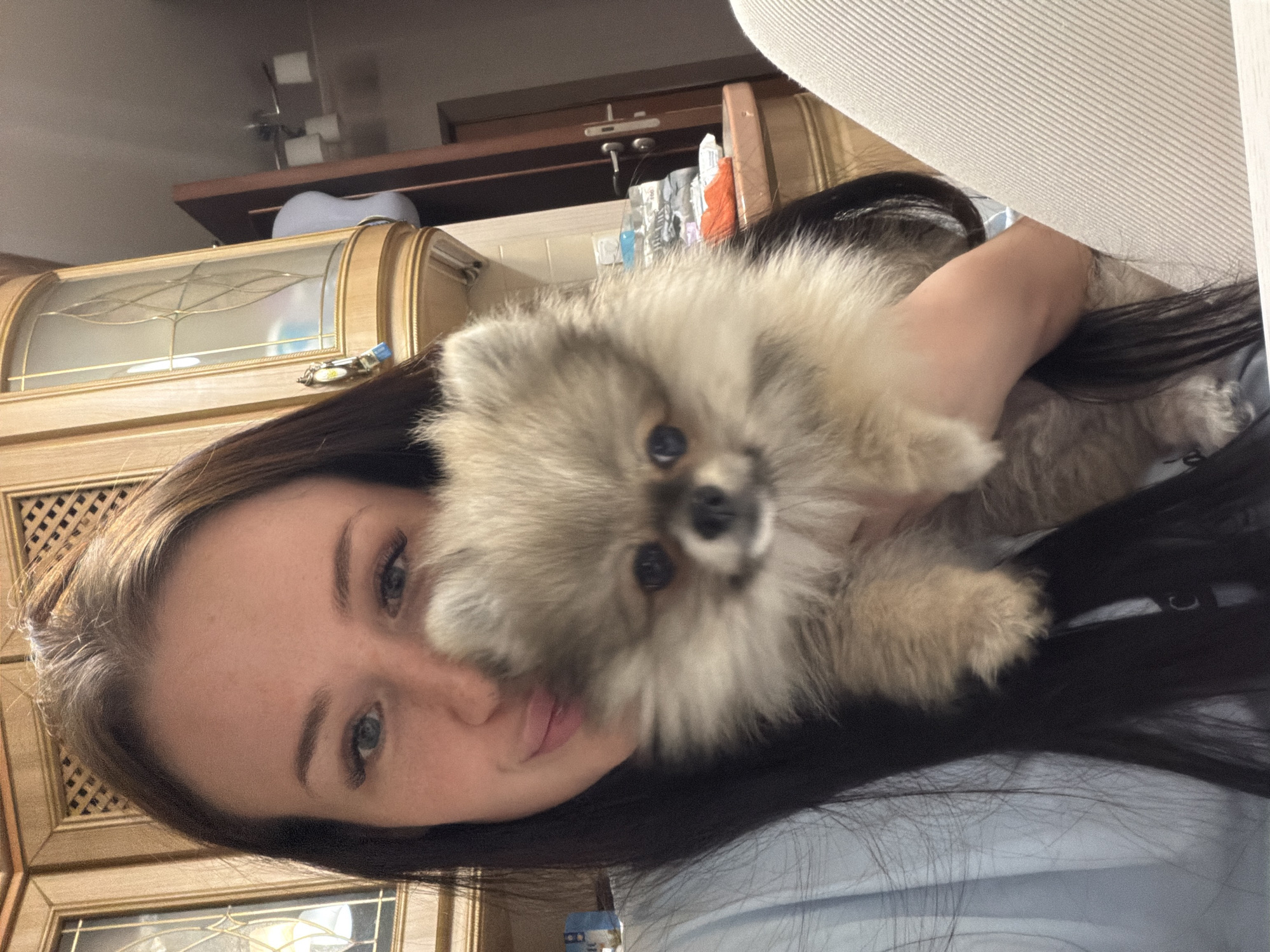 A young woman with long dark hair and light blue eyes is depicted holding a small, fluffy dog, likely a Pomeranian, close to her face in an indoor setting. The image is composed as a close-up, suggesting a selfie or an intimate moment captured. The woman wears a light blue or grey top and has a neutral to slightly smiling expression, looking directly at the camera. The dog, with its light brown and grey fur and dark eyes, is cradled in her arms, also appearing to gaze towards the viewer.
The background reveals parts of a domestic interior. Behind the woman's head, there is a wooden cabinet or hutch featuring decorative glass doors with a subtle lattice pattern. A small keychain with a blue tag hangs from one of the cabinet's handles. Above the cabinet, a darker wooden shelf holds various indistinct items, including what appears to be a grey, textured cushion or headrest. To the upper right, a light-colored, ribbed fabric, possibly part of an upholstered furniture piece or a curtain, is visible. The lighting suggests an indoor environment, possibly artificial, indicating it could be evening or daytime with drawn curtains. The scene conveys a sense of warmth and affection between the woman and her pet. The specific location is unknown, though contextually provided as Russia. No discernible text is visible in the image.