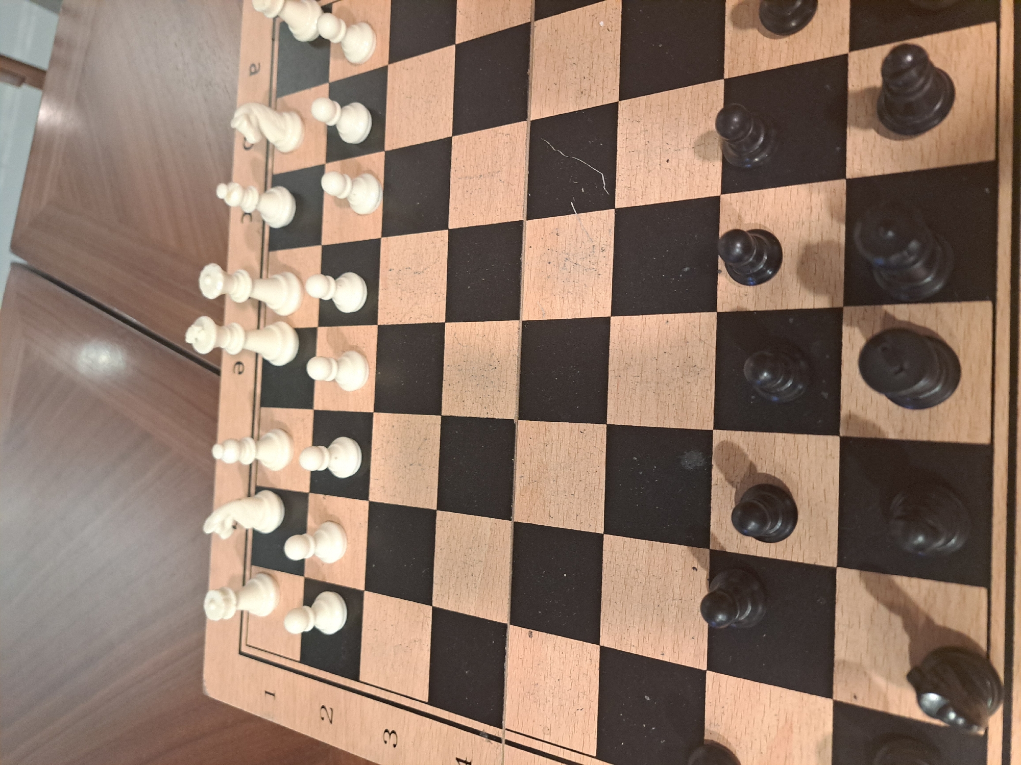 A chessboard is set up on a wooden table with white pieces lined up along the 'a' file and the first three ranks. Black pawns are lined up along the 'h' file and the sixth and seventh ranks. The location is undetermined.