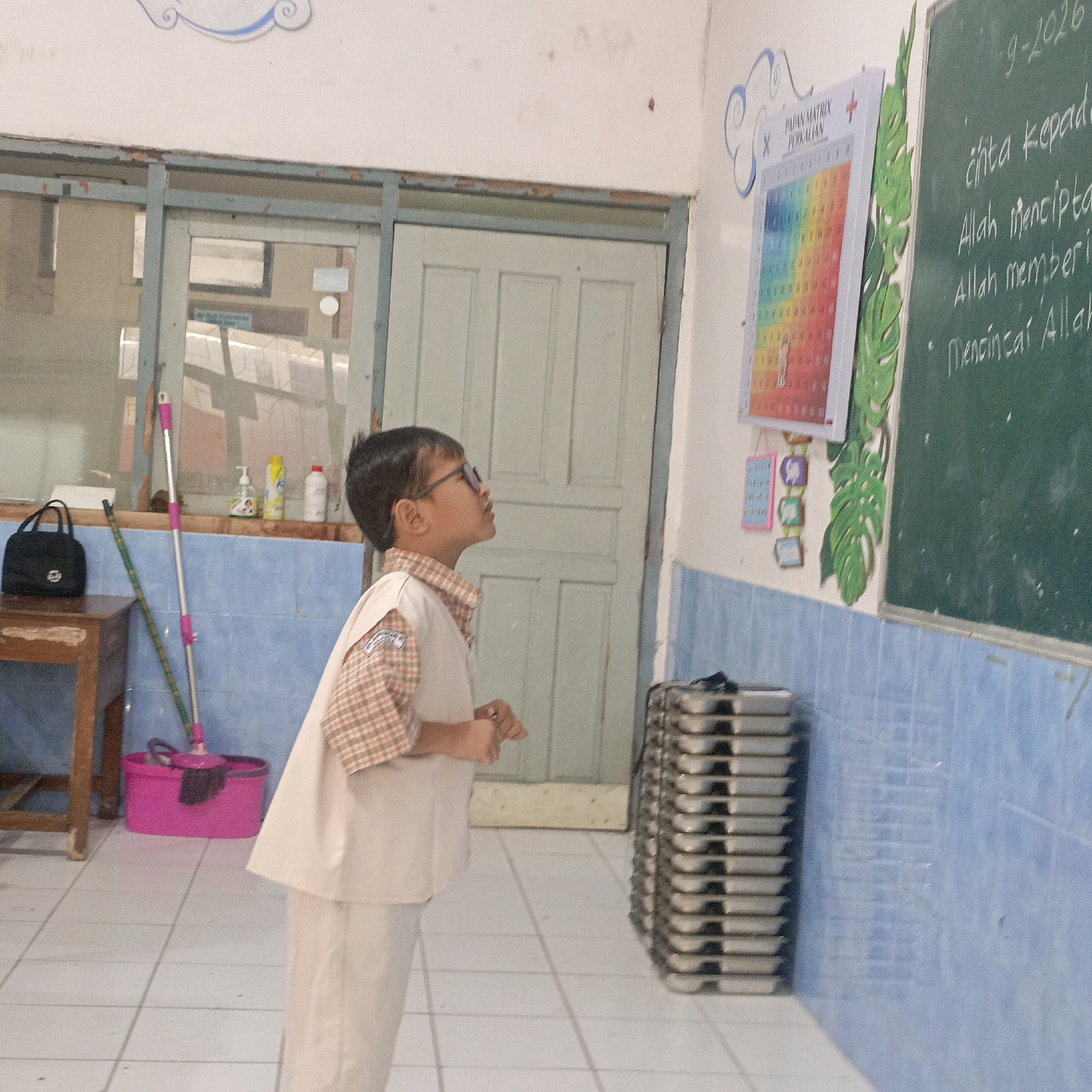 The image shows a young boy wearing glasses and a school uniform, standing in what appears to be a classroom in Salatiga, Indonesia. He is looking up at a multiplication table chart on the wall. The classroom has light blue tiled walls and white tiled floors. To the left, there is a wooden desk with a black bag and cleaning supplies, including a mop and bucket. To the right, a green chalkboard displays some writing in chalk and a decorative plant. In the background, a window and a closed door are visible. The overall impression is that of a simple, functional educational environment.