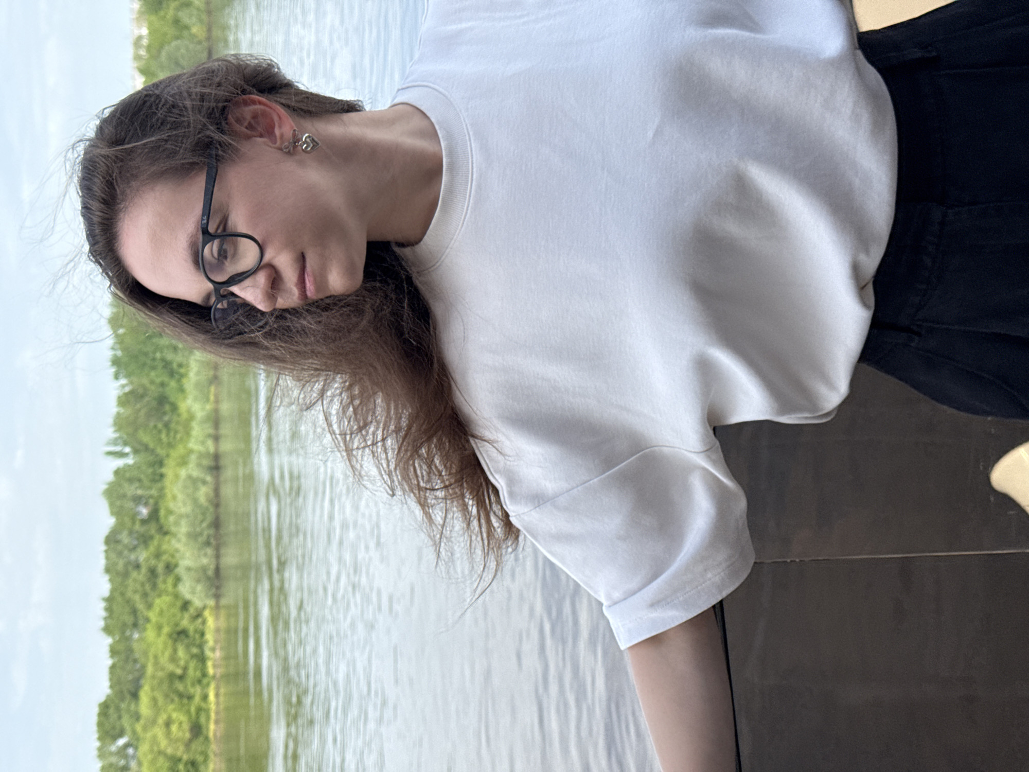 A woman with long brown hair and glasses stands outdoors, wearing a white t-shirt and black pants, near a body of water lined with green trees. The image was taken in Nizhny Novgorod, Russia.