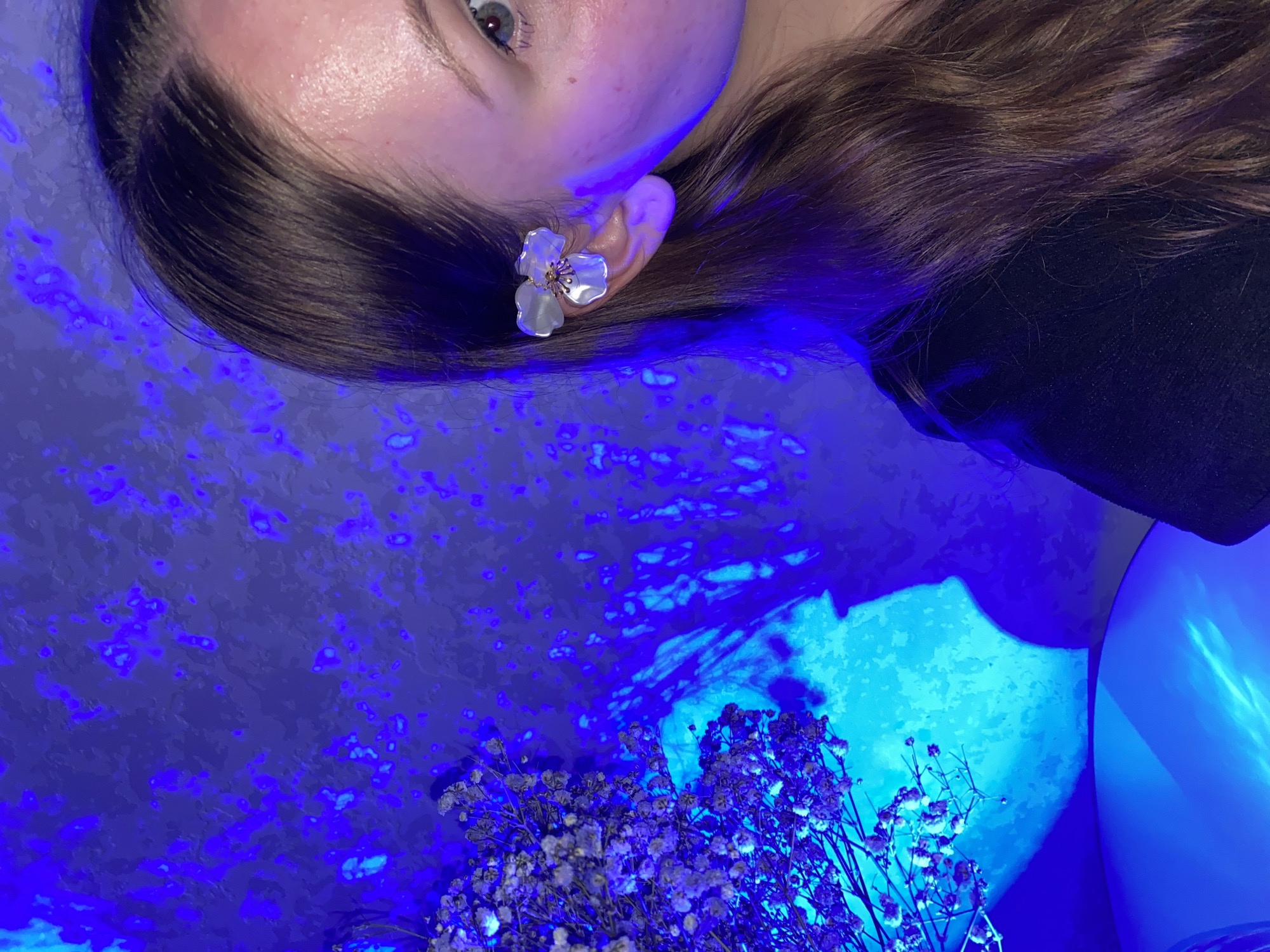 A close-up, tilted shot shows a person's face, hair, and ear with an earring, along with flowers and background lighting. The person has brown hair and an earring shaped like a white flower. The background features a mix of textures, including what appears to be a wall or surface with blue and purple lighting that creates a mottled effect. In the lower part of the frame, a bouquet of small white flowers is visible. The lighting creates strong shadows and emphasizes the textures in the scene. The location is not discernible from this image and therefore can not be related to the city of Izhevsk.