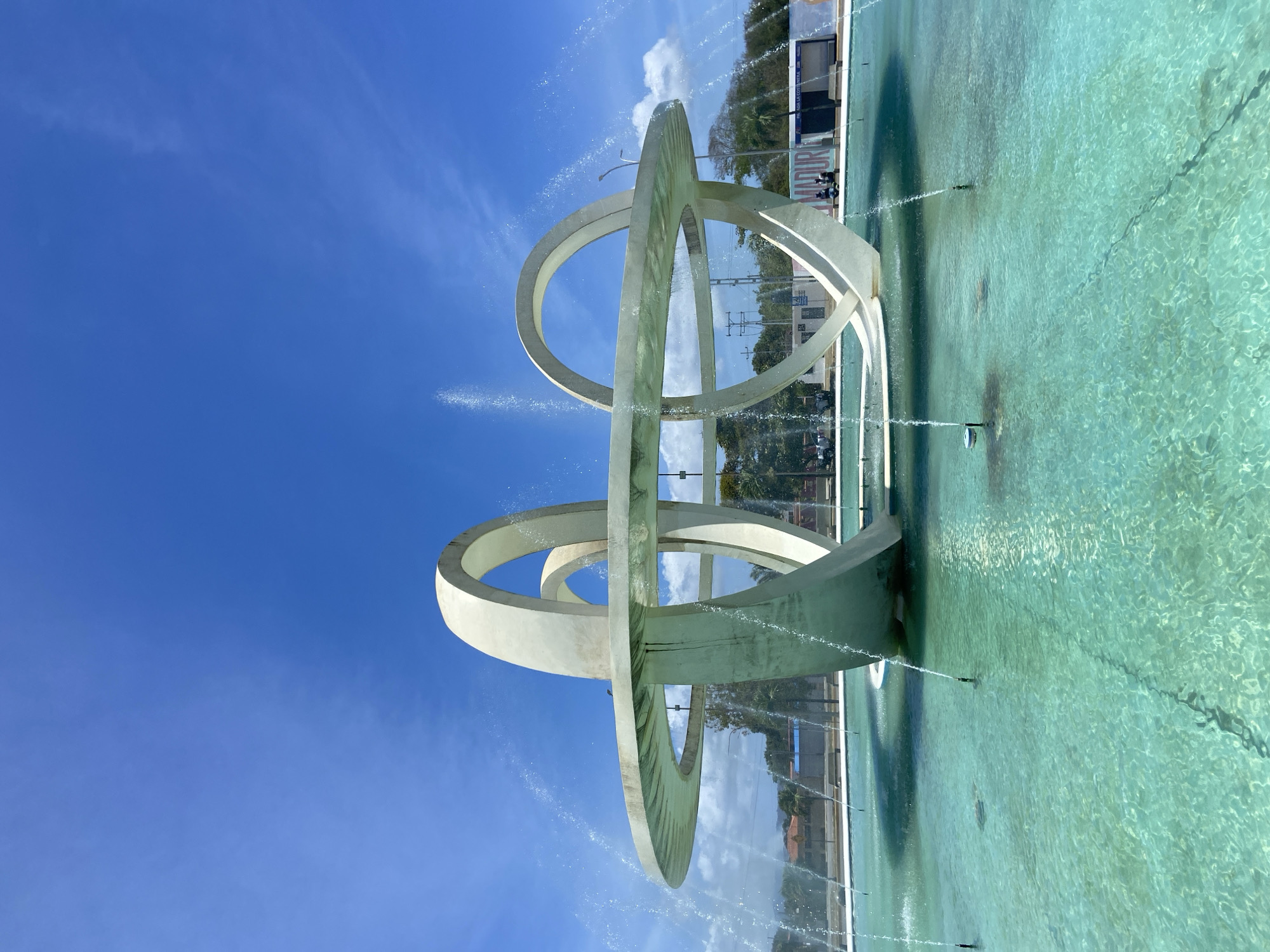 This image captures a large, active public fountain under a clear blue sky in Higuerote, Venezuela. The central subject is a monumental, abstract sculpture constructed from thick, off-white concrete elements, featuring multiple interlocking or overlapping rings. A prominent horizontal ring forms an elevated platform, from which water cascades into a circular pool below. Several vertical and diagonal ring structures provide support and define the sculpture's form within the pool. Numerous water jets actively spray upwards from the turquoise-colored water in the pool.
The environment is illuminated by strong daylight, with a bright blue sky exhibiting scattered white cumulus clouds. In the mid-ground and background, various green trees and foliage are visible, including palm trees on the right side. Behind the fountain, urban infrastructure is present, including low-rise buildings, utility poles, and an extensive network of power lines. On the far right, a structure with a large sign or mural is visible, displaying text that includes "MADURO" and "LA COSTANERA." Two individuals are seated on a concrete bench in front of this sign. A white vehicle and an additional partially visible individual are present in the left background, near what appears to be a street or roadway. The scene suggests a public plaza or roundabout.