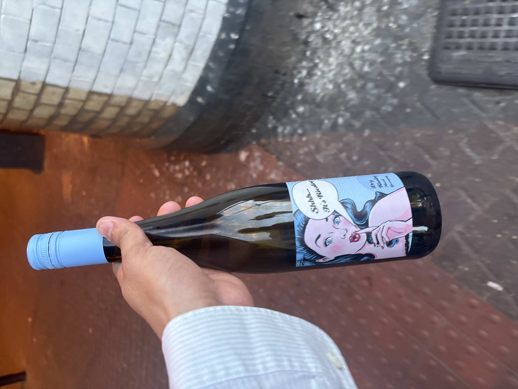 A person is holding a bottle of white wine with a light blue cap in their right hand, which is visible from the wrist down, wearing a white and light blue striped shirt. The bottle has a label with a cartoon image of a woman in a comic book style. The label reads "Shhh...It's Riesling". The background consists of a brick wall and a brown, textured surface. The location appears to be an outdoor setting in the city of London, United Kingdom, based on the architectural elements present in the background.