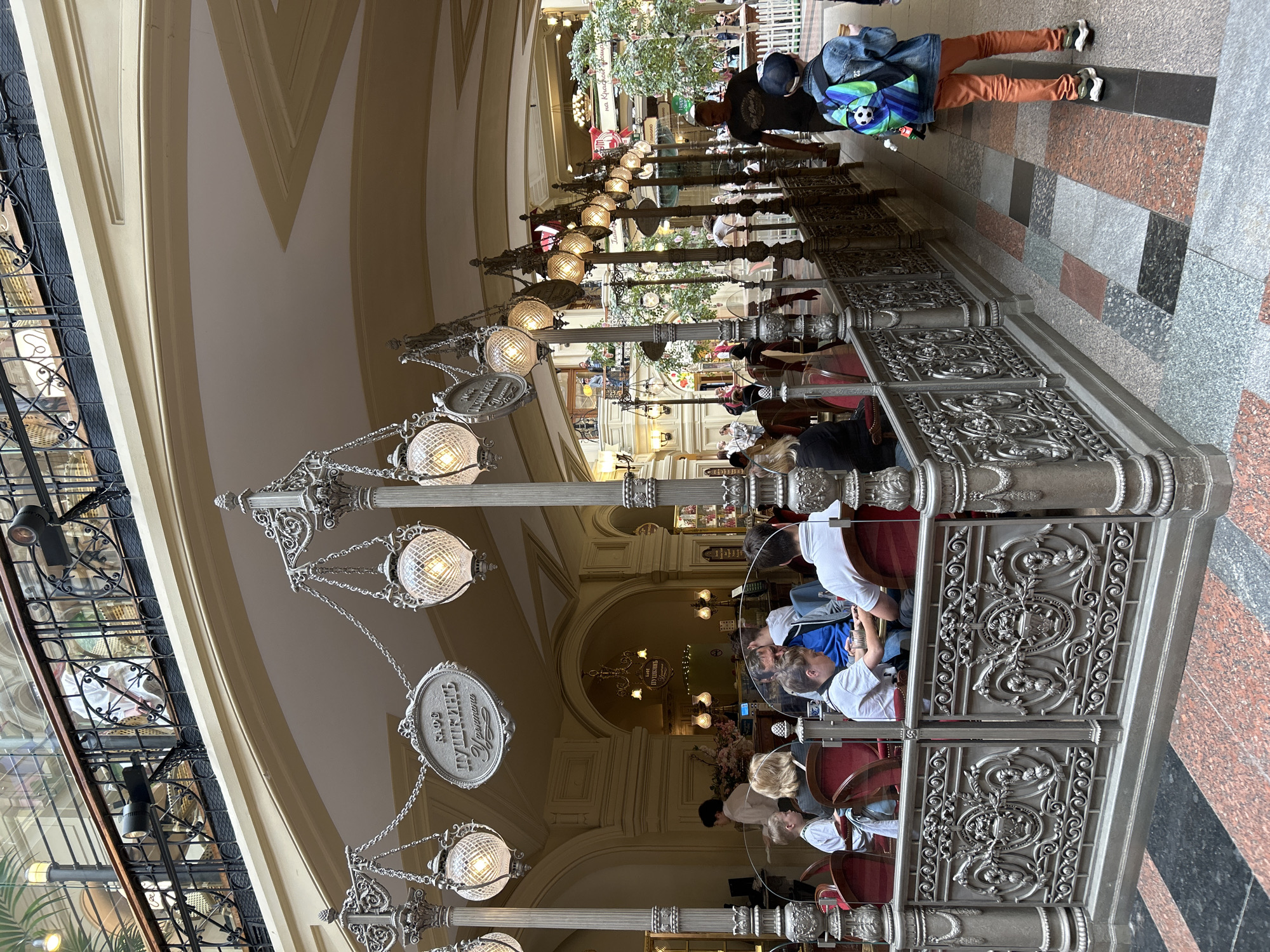 The image captures an interior view of GUM department store in Moscow, Russia, specifically focusing on an outdoor seating area of a cafe. The cafe, identified by a sign reading "КАФЕ ПУШКИНЬ" (Cafe Pushkin), is bordered by an ornate, dark grey metal railing. Several individuals are seated at tables within this area, including a family group in the foreground with men and women of various ages. The cafe's seating consists of plush red chairs.
Above the cafe, decorative lamp posts with globe-shaped lights are suspended by chains, contributing to the establishment's aesthetic. The architecture of GUM is evident in the background, featuring arched ceilings, ornate balconies with black metal railings, and multiple levels of shops. In the distance, more people are visible, as are trees and various storefronts.
To the right of the cafe, a young person wearing a denim jacket, an orange-colored shirt, and orange pants with a backpack adorned with a soccer ball is walking away from the camera. Further into the distance, other patrons are observed walking through the store. The floor is a mosaic of different colored stone tiles, predominantly granite in shades of grey, pink, and black. The overall atmosphere appears to be one of a busy, upscale shopping environment during the day.