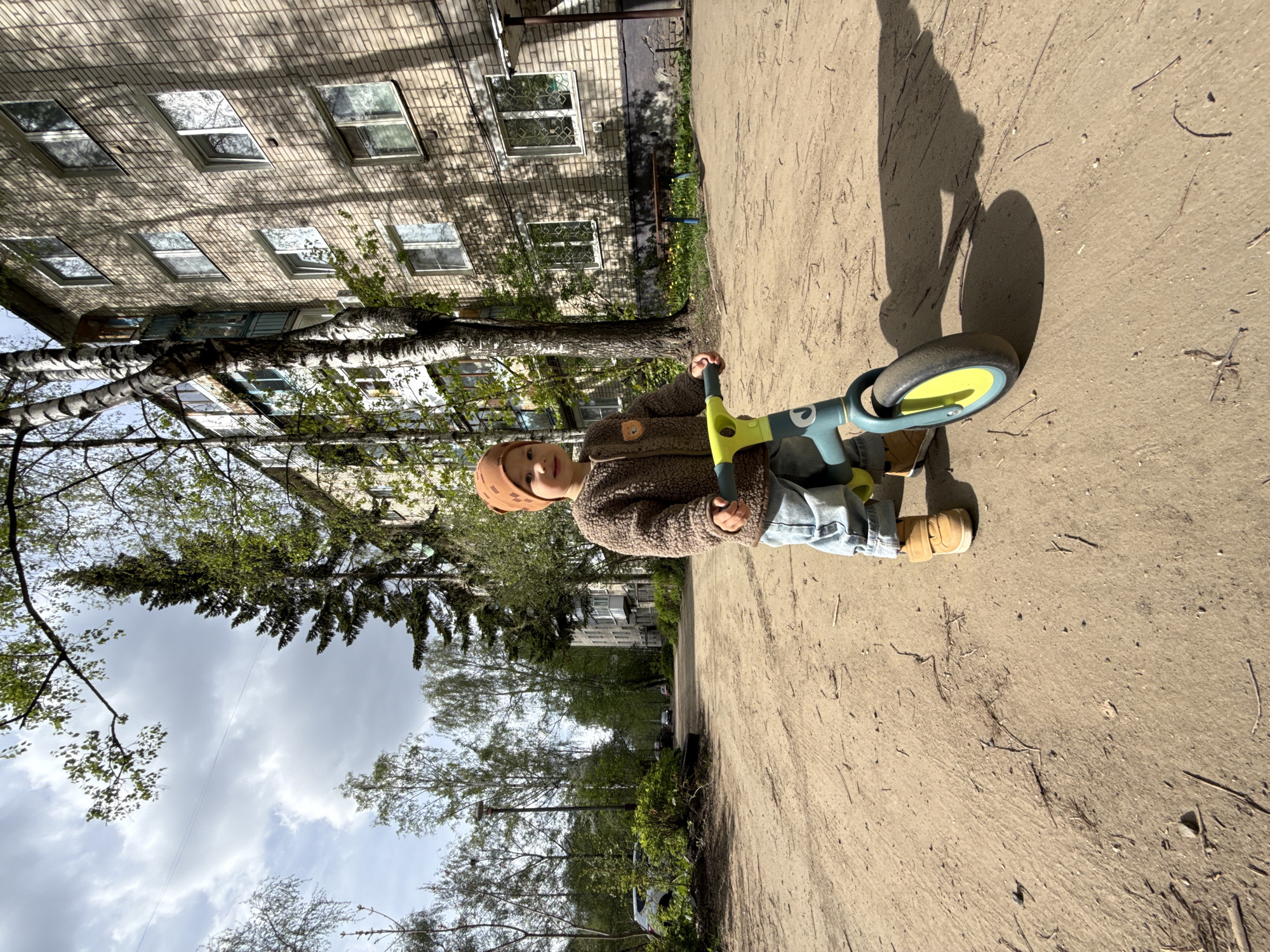 A young child in a brown hat and jacket, and blue jeans rides a light blue and yellow tricycle on a concrete path. The child's shadow is visible on the path. Behind the child is a multi-story brick building with many windows. Trees are interspersed between the building and the path. The sky is cloudy. The location is most likely Rusanovo, Russia.
