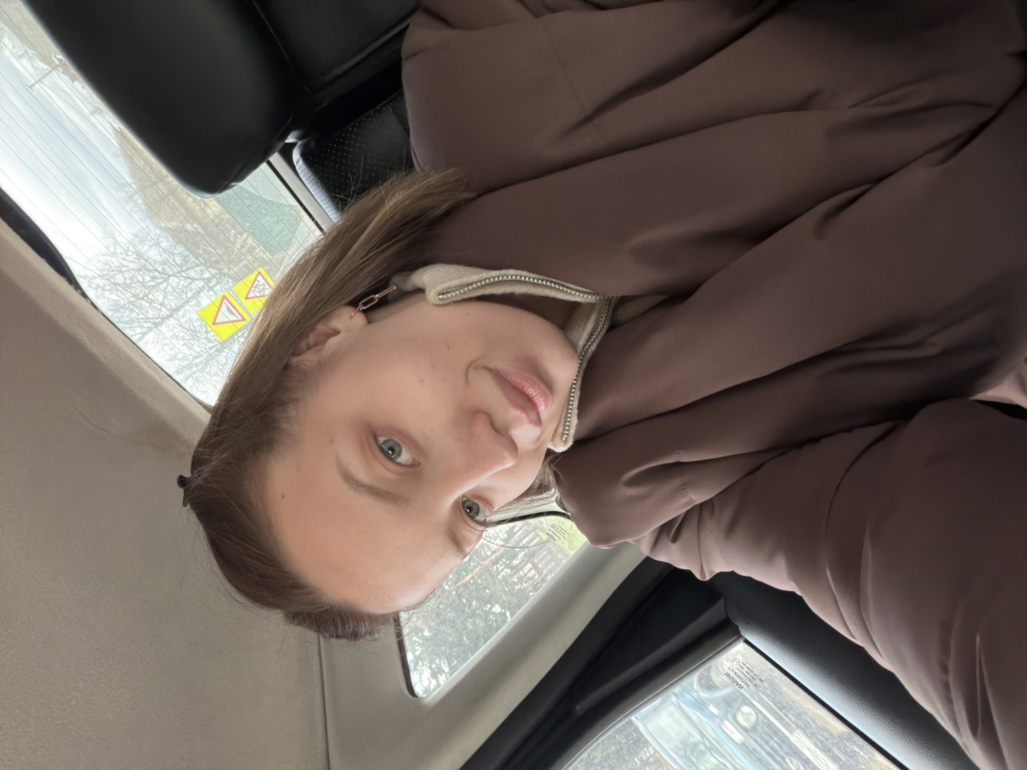 The image displays a young woman inside a car, looking directly at the camera with a calm, gentle smile. She has light brown hair and distinctive blue-green eyes. She is wearing a warm brown puffer jacket over a cream or light-colored zipped top, with the zipper partially visible. A gold-colored chain-link earring adorns her visible ear.
The setting is the interior of a vehicle, suggested by the black headrests behind her and the visible car windows. Through the car's rear window, an outdoor scene of bare trees under an overcast sky is visible, indicating a cold season (late autumn, winter, or early spring). Two yellow warning signs are affixed to the rear window; one clearly shows two opposing arrows, commonly indicating two-way traffic or a narrow road. The other sign is similar but less distinct.
The bright, natural lighting suggests it is daytime. The woman appears relaxed while taking a selfie or being photographed. Mirrored text, likely a manufacturing or safety mark (e.g., "ИОГГЭХЯУ" backwards), is visible on the side window glass. The scene depicts a moment within a vehicle in Reshetnikovo, Russia.
