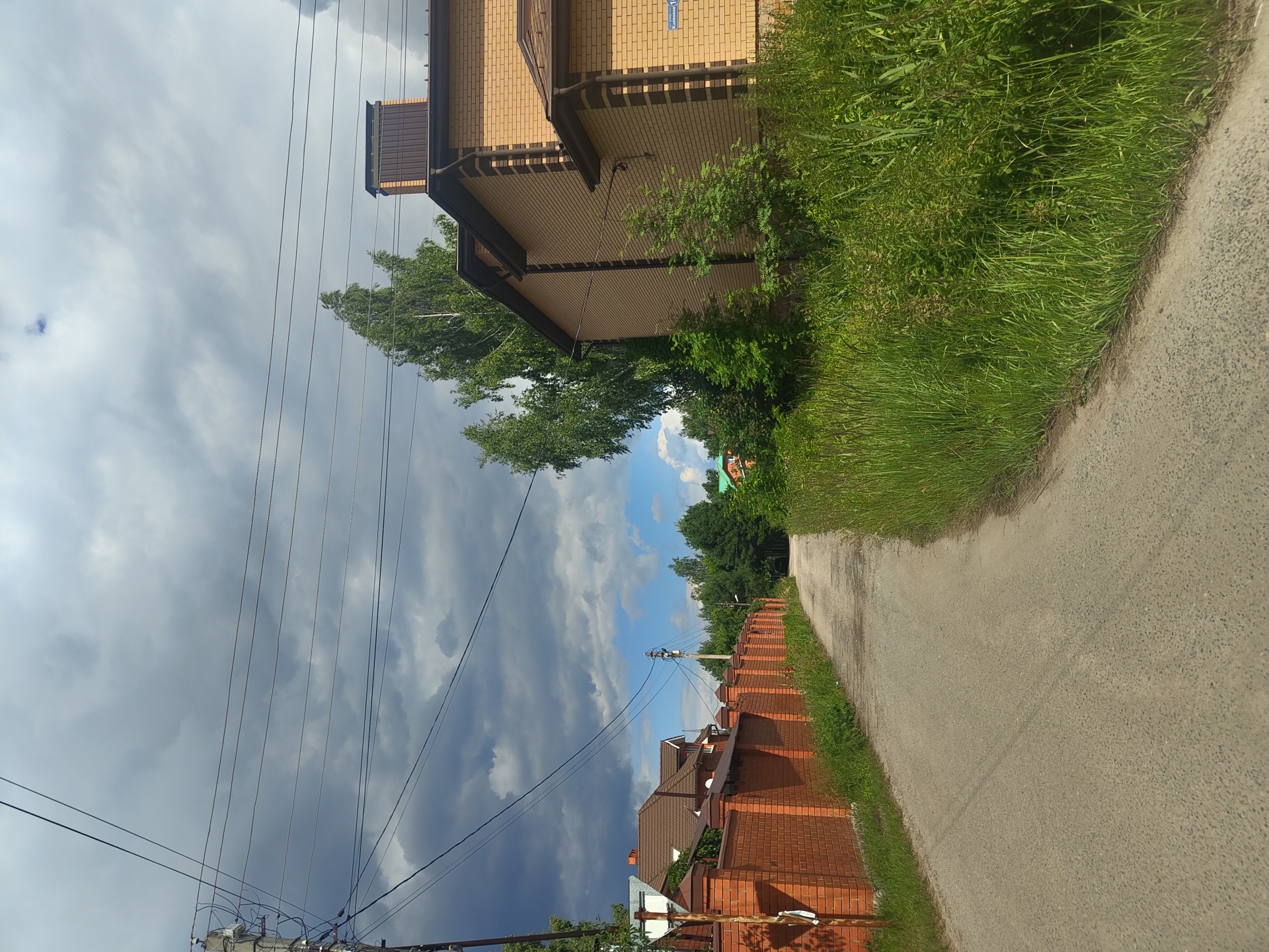 The image captures a view of a dirt road curving towards the right in the village of Tsibino, Russia. On the left, a long, red brick wall with decorative caps lines the road, separating it from a residential property with a brown-roofed house visible behind it. Overhead, power lines crisscross the sky, supported by a utility pole on the left. The right side of the road is bordered by tall, lush green grass and foliage. Further down the road, more trees and houses are visible, suggesting a suburban or rural residential area. The sky is partly cloudy, with dark storm clouds gathering in the upper left quadrant, contrasting with patches of blue sky. The scene appears to be a typical summer day, possibly before or after a rain shower.