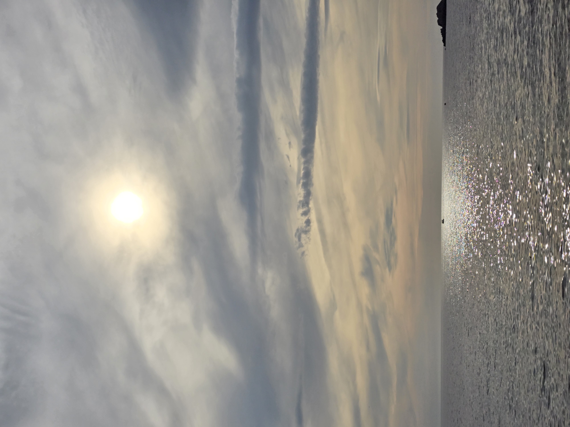 The image shows a panoramic view of the Adriatic Sea off the coast of Perazića Do, Montenegro. The sun is partially obscured by wispy clouds, casting a shimmering reflection on the water's surface. A small boat can be seen in the distance, and a rocky outcrop is visible on the right side of the frame. The sky is a mix of grey and pale yellow, with streaks of darker clouds suggesting a change in weather. The overall impression is one of a serene, yet dramatic, seascape.