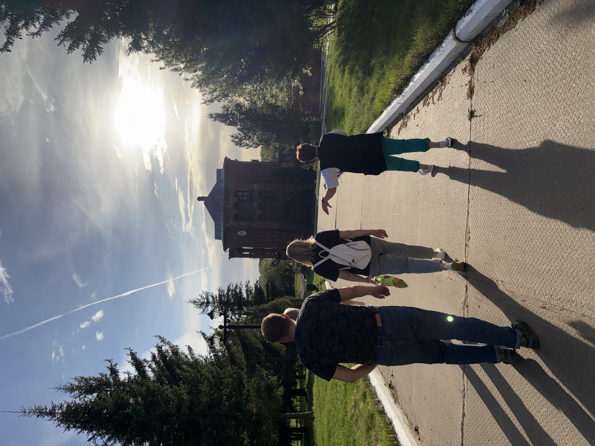 Three people are walking on a pavement next to a grassy area. The location is Zavod Mikhailovskii, Russia, as inferred from visual details. The first person, closest to the viewer, is wearing a dark patterned shirt, blue jeans, and dark shoes. The second person has blonde hair and a white backpack, wearing a dark shirt and light-colored pants. The third person wears a white shirt with a dark vest, and green pants. A historic brick building is visible in the background against a bright sky with some clouds and a contrail. Tall trees line the left side of the pavement.