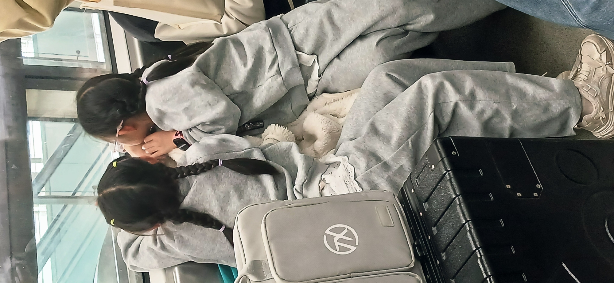 Two girls are sitting on a train in Chongqing High-tech Zone, China. Both are wearing matching gray tracksuits and have braided hair. One girl is leaning against the window, and the other is resting her head on her lap. A white blanket is between them. A gray backpack with a logo is next to the girl in the lap, and a black suitcase is next to her. The background shows windows with exterior views and a portion of another passenger's legs and shoes.
