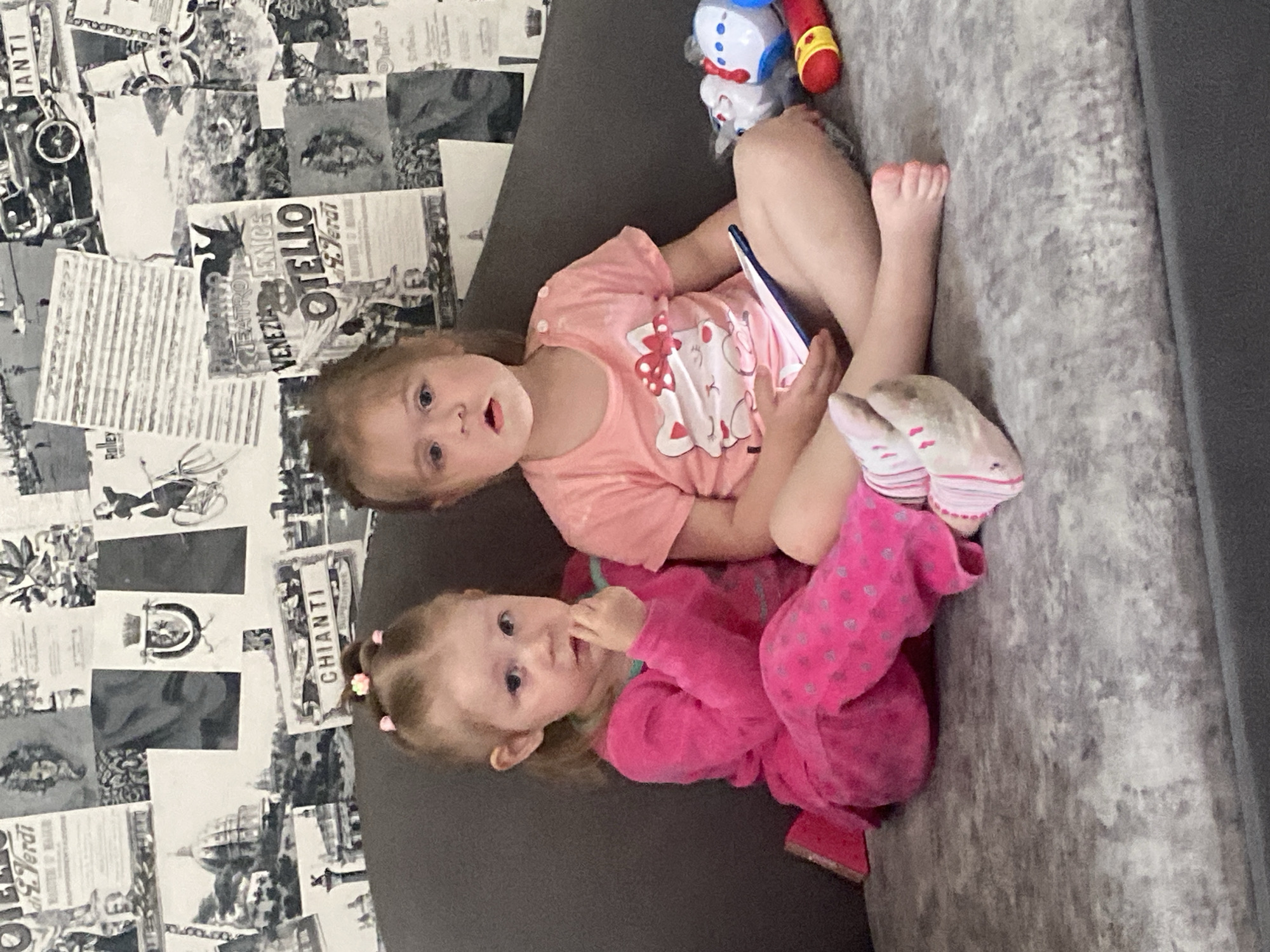 Two young girls are lying on a gray sofa. The girl on top is wearing a light pink shirt with a Minnie Mouse design and has a tablet in her hands. She is looking directly at the camera. The girl below is wearing pink pajamas and has her finger in her mouth. The background includes a black and white wallpaper featuring various Italian landmarks and advertisements. A toy is near the top of the image. The scene appears to be indoors. There is no clear indication of location in Chaikovsky, Russia.