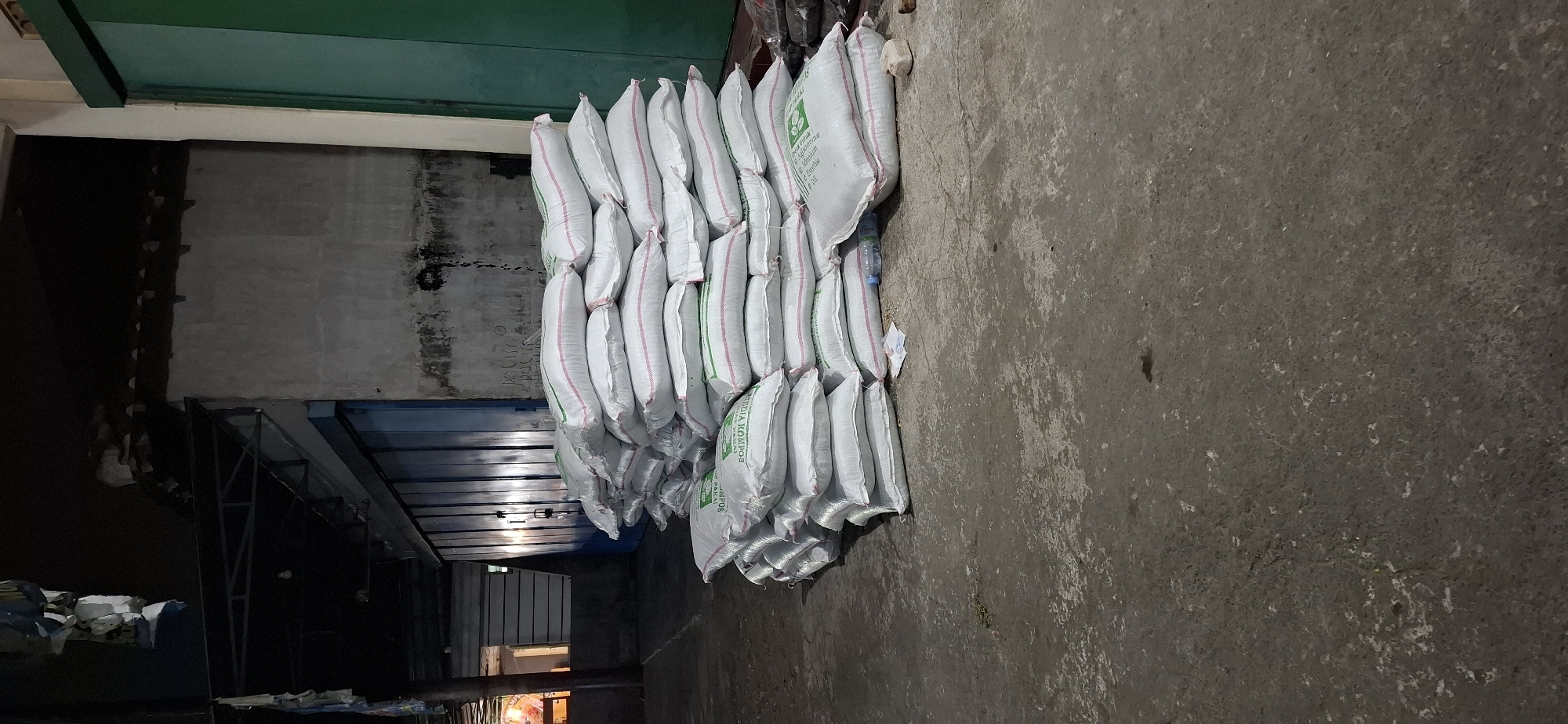 The image depicts an outdoor scene in Klumprit, Indonesia, likely at a market or storage area. A large, uneven pile of white sacks, each with red and green stripes and some with visible green lettering, dominates the foreground. The sacks are stacked haphazardly, with some appearing slightly disheveled. A portion of the sacks has "MEDIA KOMPOS" printed on them.
In the background, a building with a textured, off-white wall is visible. This wall has dark, irregular marks, possibly stains or graffiti, near its base. To the left of the building, a blue roll-up door or shutter is partially visible. Above the building, a dark, overhanging structure with metal supports can be seen. Hanging from the left side of the frame are some tattered white and blue banners.
To the right of the sacks, a dark green door or panel is present. The ground is made of concrete, showing signs of wear and discoloration. A few small stones and a clear plastic bottle are scattered on the ground near the sacks. The lighting suggests it is either dusk or night, with artificial lights illuminating the area. There are no people visible in the frame.