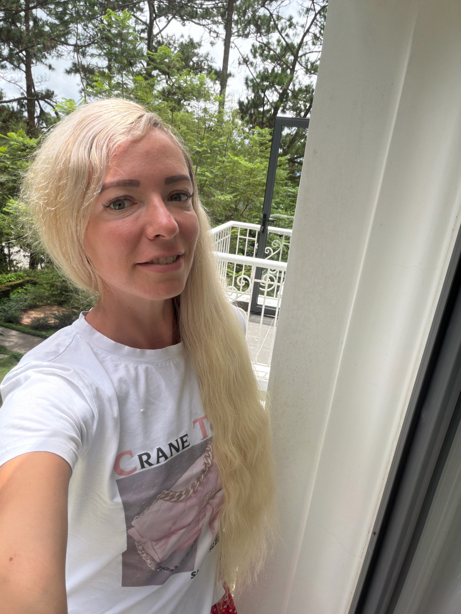 A woman with blonde hair is taking a selfie on a balcony in Xuân Trường - Đà Lạt, Vietnam. She is wearing a white t-shirt with a graphic of a pink purse and text that reads "CRANE T". Behind her, trees and a white railing are visible. The balcony appears to be part of a residential building, possibly a hotel or guesthouse. The weather is overcast.