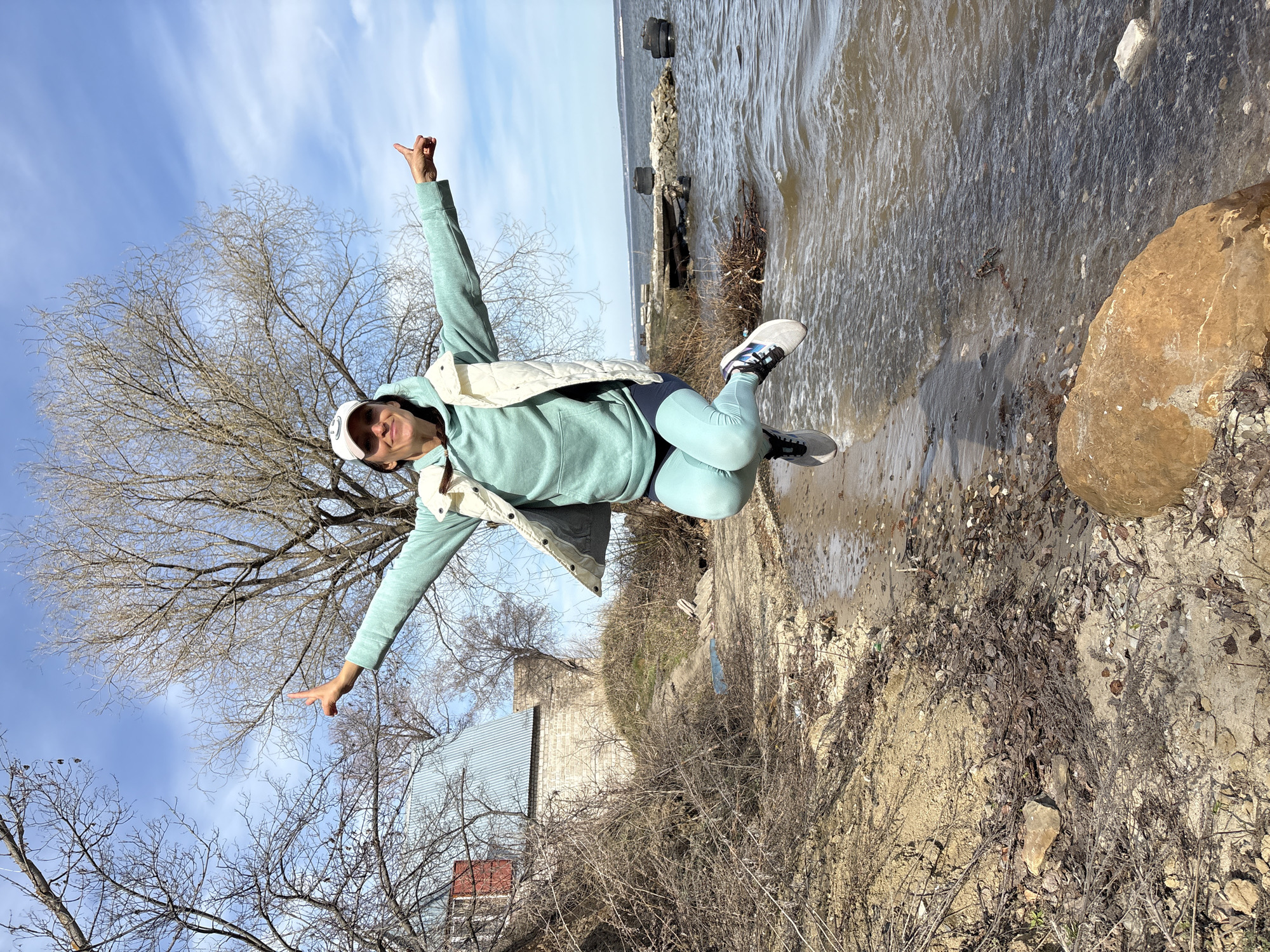 A dynamic outdoor shot captures a woman leaping mid-air with outstretched arms, expressing joy and freedom. She is dressed in a light mint green hoodie and matching leggings, a white puffer vest, a white baseball cap, and grey and white athletic shoes with blue accents. Her hands are open, one making a gesture resembling the 'hang loose' sign, and her face shows a happy expression.
The scene depicts a shoreline or riverbank in Ulyanovsk, Russia, likely during late winter or early spring, given the bare branches of the trees and the attire. The background features a wide body of water, possibly the Volga River, with visible concrete structures or a pier near the water's edge. The water appears somewhat murky and choppy. On the left side of the frame, several large deciduous trees with intricate, leafless branches stand against a bright blue sky with scattered wispy clouds. Further back on the left, a light-colored brick building with a dark corrugated metal roof and a smaller red structure are visible through the trees. The ground is a mix of dirt, dry grass, and sparse, dead vegetation, with some large rocks scattered near the water. The overall impression is one of energetic movement against a somewhat rugged natural and semi-urban backdrop on a bright, clear day. There is no visible text.