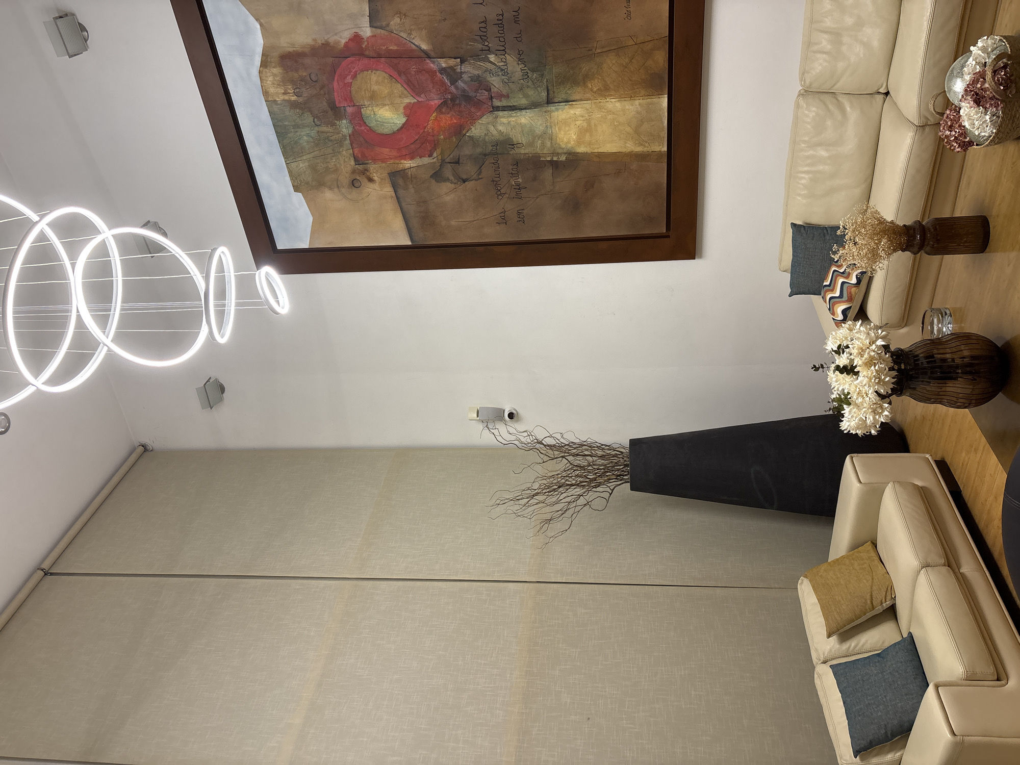 This media file captures a contemporary living room or lounge area, viewed from a slightly elevated perspective. The setting suggests a modern indoor space, potentially a residential home or a hotel common area, consistent with a location like San Bernardino Tlaxcalancingo, Mexico.
The scene is dominated by two light-colored, possibly cream leather sofas, arranged at an angle to each other. One is a longer sofa, and the other a shorter one, both adorned with decorative throw pillows, including one solid blue, one mustard yellow, and one patterned with blue, brown, and orange chevrons. A wooden coffee table is partially visible between them.
A large, abstract painting in a dark brown frame hangs on the wall above the longer sofa. Its palette features earthy tones of brown, gold, and red, with circular and organic shapes. Handwritten Spanish text is visible on the artwork, including phrases like "todas las posibilidades dentro de mi" (all the possibilities within me) and "las oportunidades son infinitas y" (the opportunities are infinite and).
The room features bright, modern artificial lighting from a unique chandelier composed of multiple glowing white LED rings, arranged in a cascading and overlapping pattern. Two wall-mounted speakers are visible, as well as a small security camera or sensor mounted higher on the wall.
On the coffee table, various decorative items are arranged: a dark brown ribbed vase with dried white flowers, another dark brown vase holding fine beige dried grasses, and a decorative basket containing artificial pink and white flowers. A tall, dark-colored floor vase filled with dried, slender branching sticks stands prominently near the shorter sofa.
The far wall is covered by light beige roller blinds or shades, suggesting large windows behind them. No people are present, and no specific activity is taking place. The overall impression is one of modern elegance and calm. The bright artificial light suggests it could be evening or night, or a heavily shaded daytime interior.