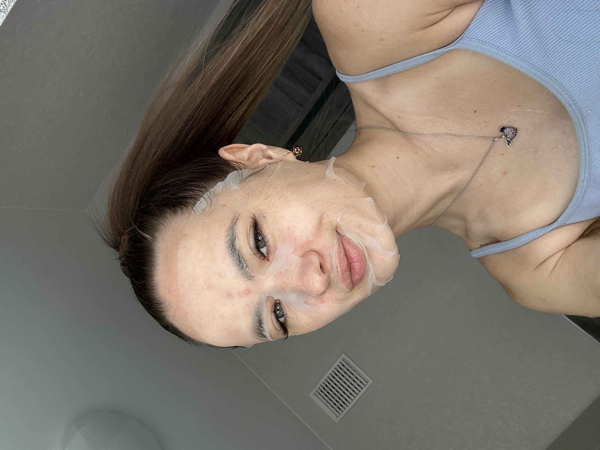A young woman with a high ponytail and a sheet mask on her face is taking a selfie in an indoor environment. She is wearing a light blue tank top and a silver necklace with a heart-shaped pendant. Her makeup includes false eyelashes and mascara. The background is a plain gray wall with a white vent. The lighting appears to be natural, coming from above.