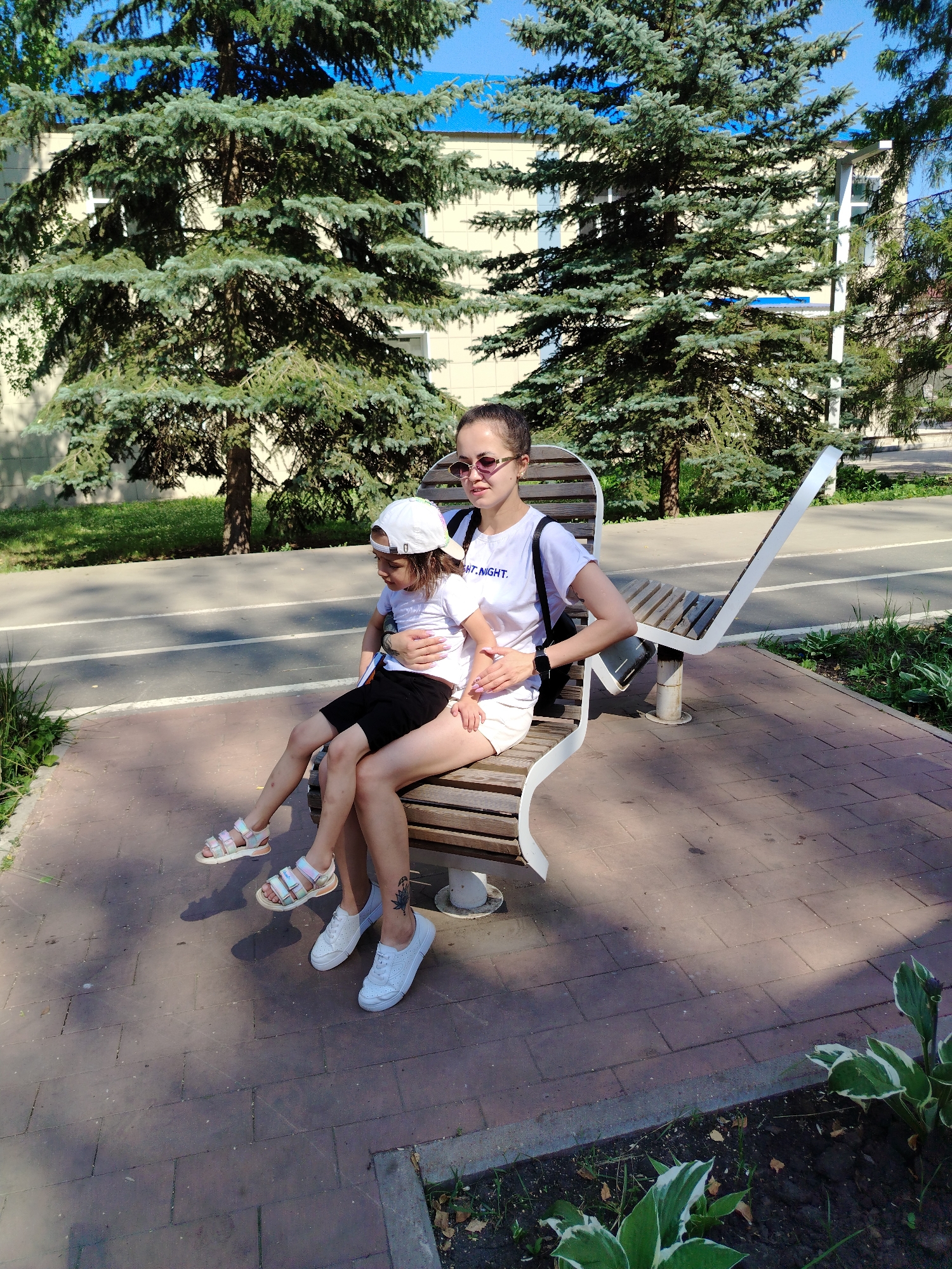 The image depicts two individuals, likely a mother and child, seated on a modern, curved park bench. The woman, positioned on the right, is wearing a white t-shirt with blue lettering and white shorts. She has her arm around the child, who is seated on her lap. The child is wearing a white t-shirt, black shorts, and a white baseball cap. Both individuals are wearing casual footwear. The bench is situated on a brick-paved pathway, bordered by a flowerbed with green and white variegated plants. Tall evergreen trees provide a backdrop, with a building visible behind them. A paved area, possibly a bike path, is visible in the middle ground. The scene appears to be a public park in Yanaul, Russia, on a sunny day.