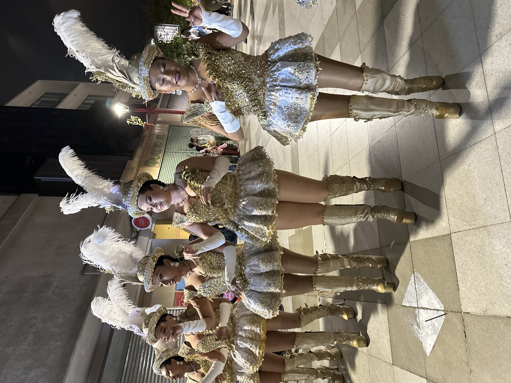 In Arica, Chile, a group of women in elaborate, sequined gold and white costumes with feathered headdresses are performing. They are wearing knee-high, platform boots. The performance appears to be outdoors, possibly on a street or plaza, with paved ground tiles. In the background, there are buildings and streetlights, suggesting it is nighttime. Some of the women are looking towards the camera, and one is making a peace sign. The overall scene suggests a festive or celebratory event.