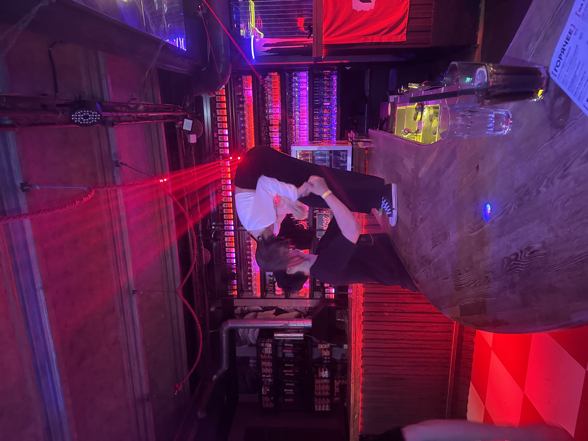 A person in a white shirt and black pants is bending over at a bar. Another person in a black t-shirt is standing next to them. The bar is lit with red and purple lights. The floor is a red and white checkered pattern. There are bottles of alcohol on shelves behind the bar. The scene appears to be in a nightclub or bar.