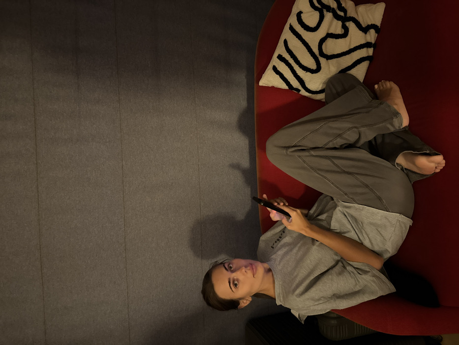 A female subject is depicted reclining on a red upholstered sofa in an interior, dimly lit setting. She has long, dark hair, pulled back, and is wearing a grey, short-sleeved t-shirt and grey sweatpants; her feet are bare. She holds a black smartphone with both hands, positioned over her lap, and appears to be interacting with the device. Her gaze is directed forward, towards the viewer. To her right, on the sofa, a cream-colored square throw pillow with a black abstract wavy line pattern is present. The wall behind the sofa is grey and features horizontal ridges, consistent with acoustic paneling or textured wallpaper. Shadows are cast on this wall, notably behind the subject's head and upper torso, indicating an off-camera light source. There are no discernible visual cues within the image to identify a specific location in the city of Warsaw, Poland.