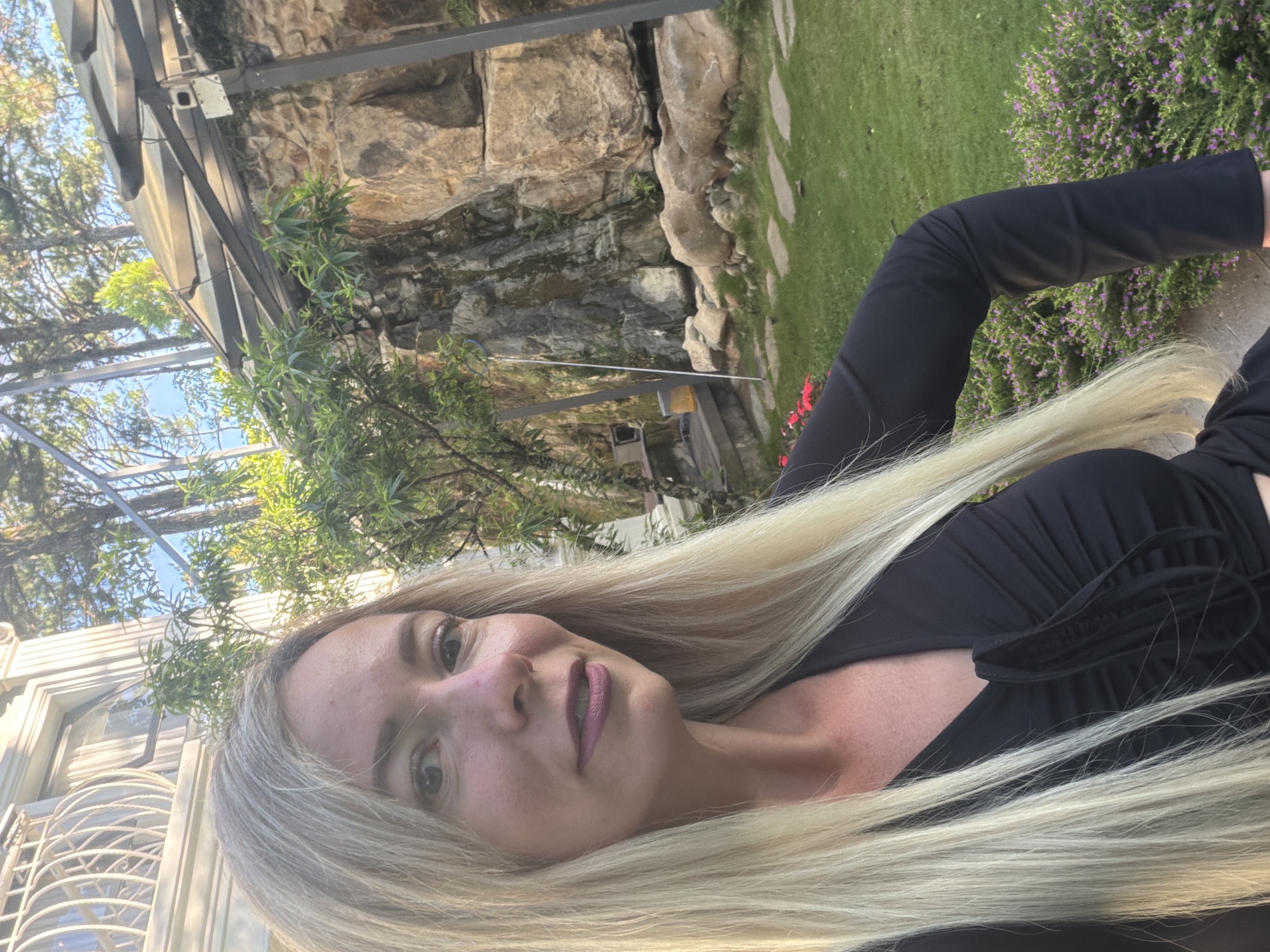 A blonde woman with long hair and dark clothing is captured in a selfie outdoors. She is positioned in the foreground, with her upper body facing the camera. Her expression is a slight smile with her lips slightly parted. Behind her, a stone wall with some greenery and trees is visible. The scene appears to be in a garden or outdoor recreational area. The location is identified as Xuân Trường - Đà Lạt, Vietnam.