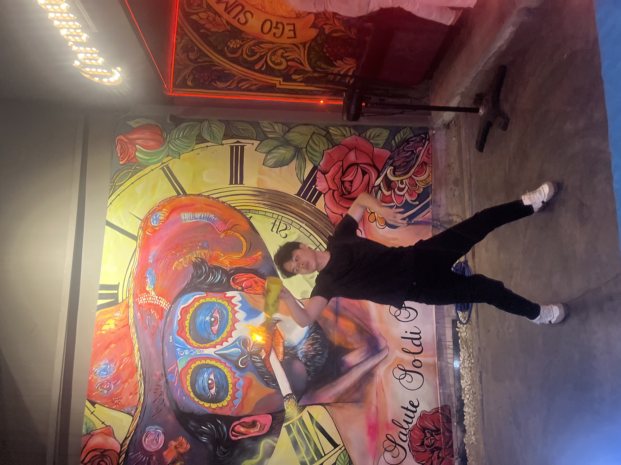 The image captures a young man performing a trick with a lit cigarette, possibly fire breathing or juggling, in front of a vibrant, large-scale mural in Đà Nẵng, Vietnam. The mural depicts a stylized portrait of a man with Day of the Dead-inspired face paint, wearing a wide-brimmed hat adorned with colorful patterns and roses. The background of the mural features elements of a clock face and more roses. To the right of the mural, a neon sign, partially visible, spells out "Corona." The man is dressed in a black t-shirt and black pants, and he is mid-action, with his right arm extended and holding a lit cigarette that is aflame. His left arm is bent at the elbow, with his hand positioned near his chest. He is wearing white and grey sneakers. The floor is a concrete surface, and there are some rocks and a blue hoop visible in the foreground. The lighting suggests an indoor or partially covered outdoor space, possibly a bar or entertainment venue, with red neon lighting accentuating the right side of the scene. The mural is the dominant visual element, creating a dramatic and artistic backdrop for the man's performance.