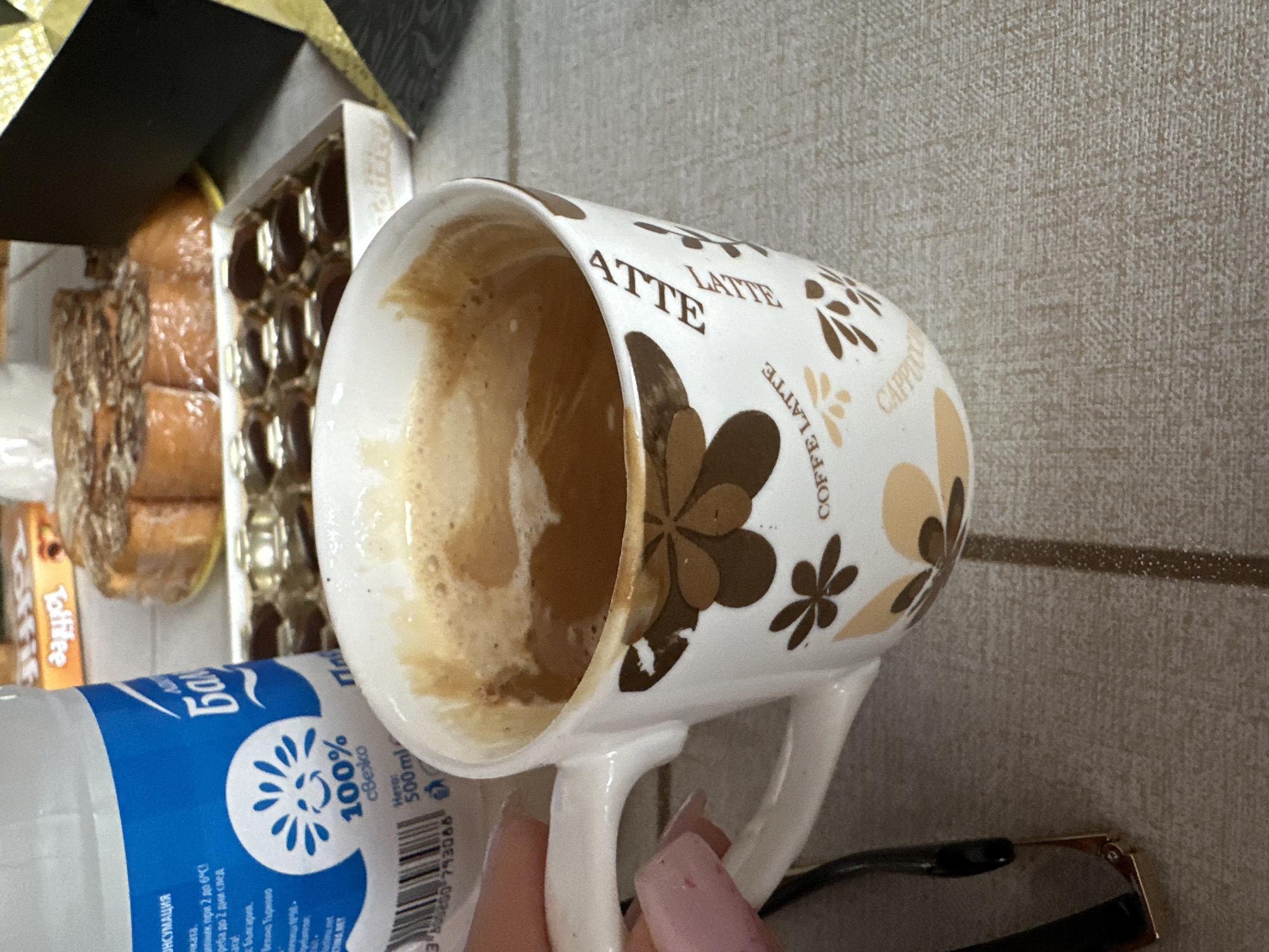 The image shows a partially consumed latte in a white mug with brown and beige floral designs and the words "LATTE" and "COFFEE LATTE" printed on it. Next to the mug is a blue and white bottle with Cyrillic text and a symbol that reads "100% свежо" (100% fresh). There is also a package of sweets, possibly chocolates or similar treats. A finger with a pink nail is holding the mug. The background features a neutral-toned, textured surface. A pair of glasses is also visible in the bottom right corner. There is not enough information to determine the location in Montana, Bulgaria.