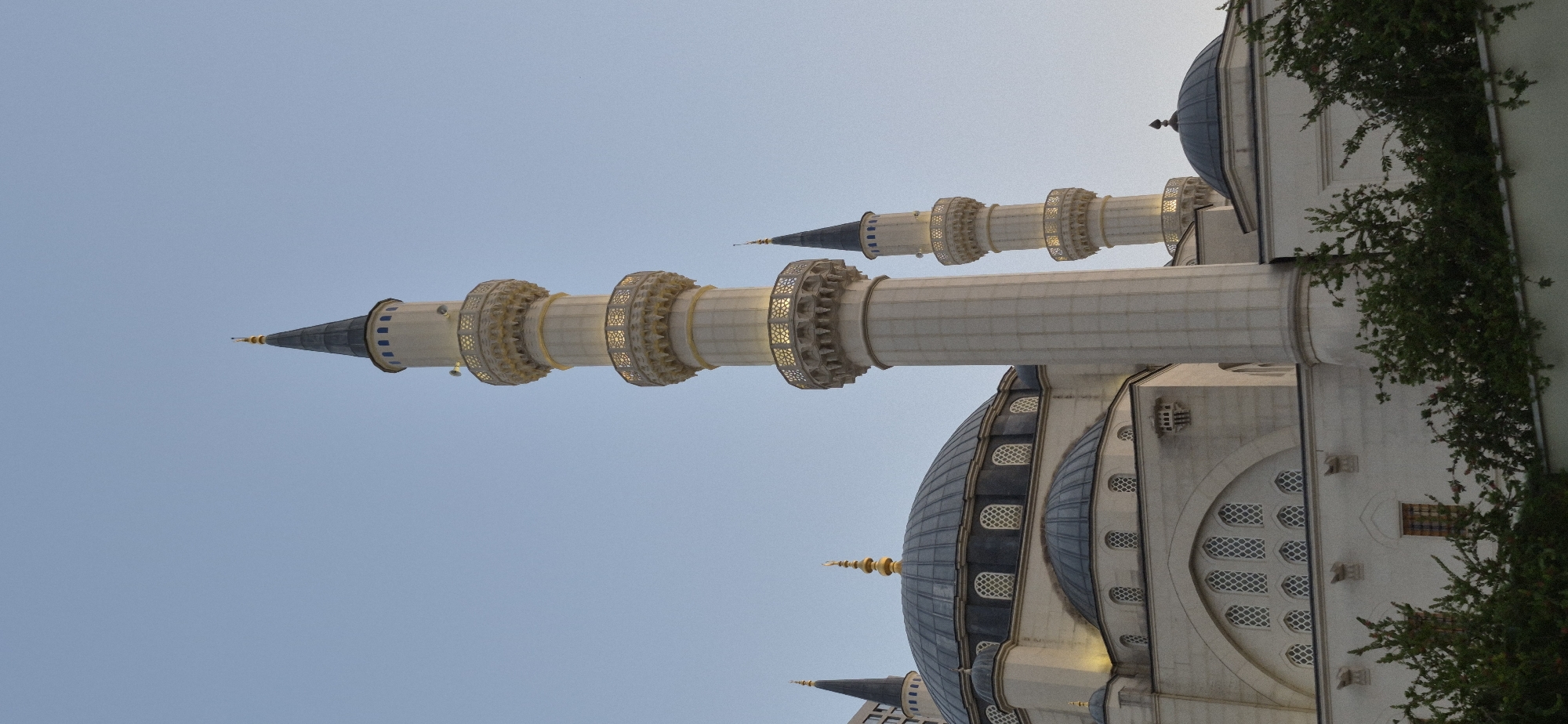 The image shows the exterior of the Et'hem Bey Mosque in Tirana, Albania. The mosque features a large central dome and several minarets. The architecture is light-colored stone with ornate details on the minaret balconies and dome spires. The sky is a clear, pale blue, suggesting it is daytime, possibly early morning or late afternoon. Green foliage is visible in the foreground, obscuring the lower part of the mosque's facade and a light-colored wall. The overall scene is peaceful and architectural.