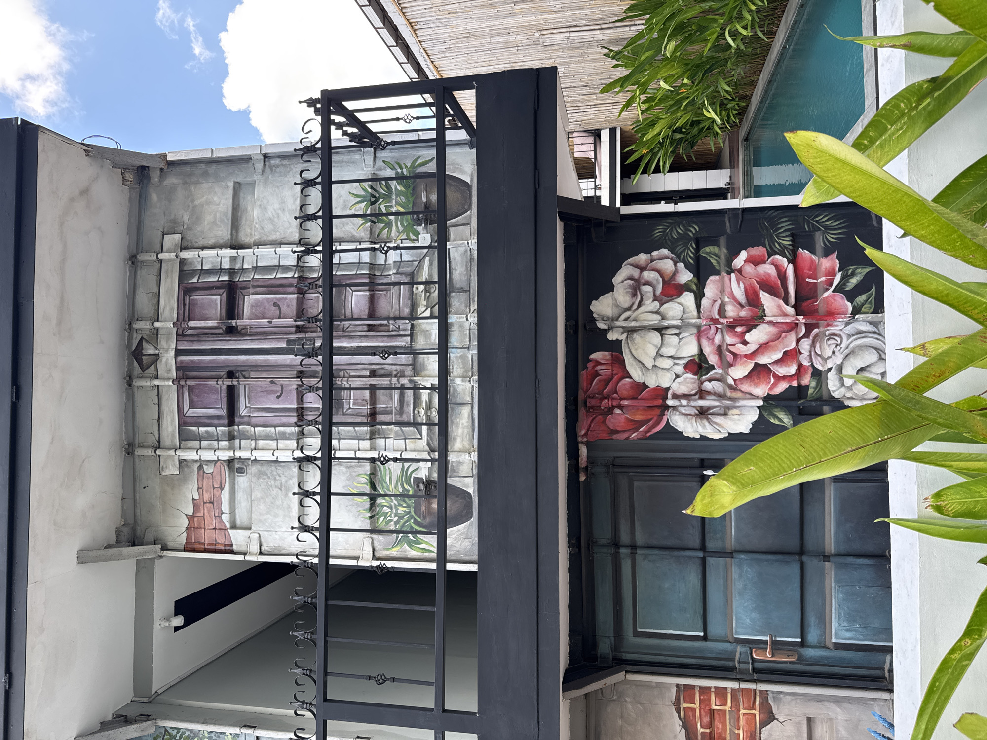 An exterior shot of a multi-story building in North Kuta, Indonesia, features artistic murals and a balcony overlooking a swimming pool. The upper portion of the building displays a painted facade resembling an old European-style window with dark purple shutters and decorative ironwork. Two potted plants flank the window mural. A black metal railing encloses a balcony on the second level. Below the balcony, the building's facade is adorned with a vibrant mural of large, detailed pink and white roses painted against a black background. To the right of the rose mural, a narrow swimming pool with clear blue water is visible, surrounded by lush green foliage. The sky above is a bright blue with scattered white clouds. The building appears to be a hotel or resort, given its aesthetic and the presence of the swimming pool.