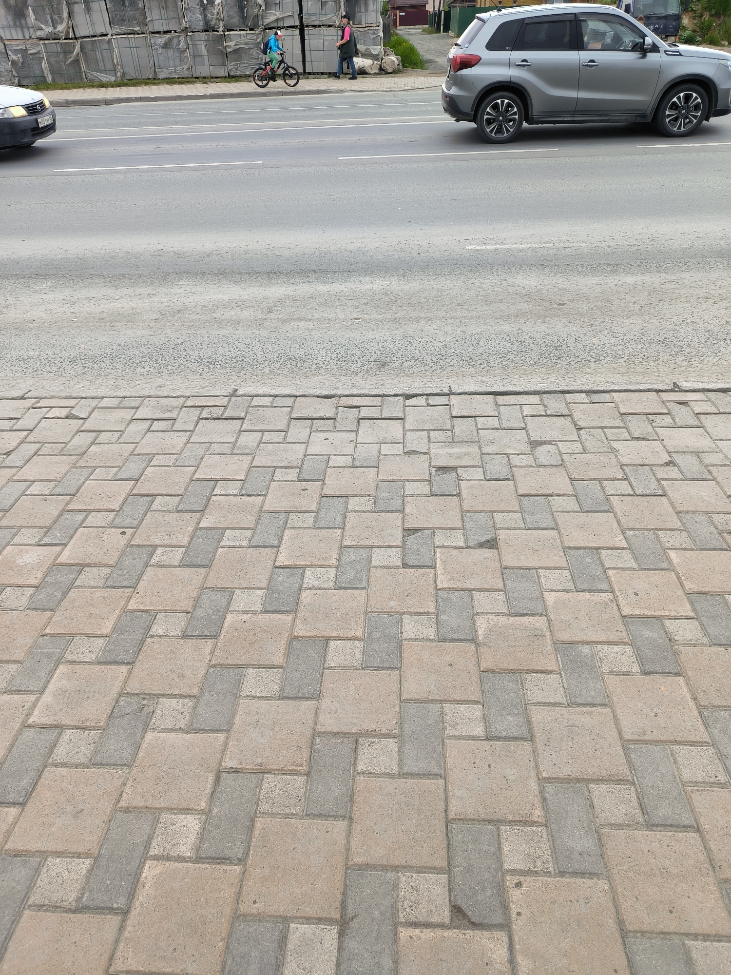 The image depicts a street scene in Yuzhno-Sakhalinsk, Russia. In the foreground, a paved sidewalk is visible, composed of rectangular and square interlocking pavers in alternating shades of tan and gray. The sidewalk leads towards a road. On the road, a silver SUV is driving in the right lane, and a white car is in the left lane. Further down the road, a person on a bicycle, wearing a blue jacket and a red cap, is riding in the same direction as the SUV. Another person in a pink jacket is walking on the sidewalk near the cyclist. In the background, stacks of gray materials, possibly building supplies, are visible along the side of the road, along with some buildings and vegetation. The sky is overcast, suggesting a cloudy day.