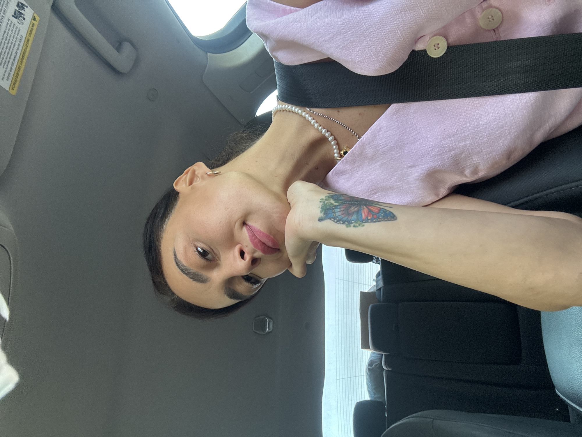 A woman with dark hair is in the passenger seat of a car, looking at the camera and resting her chin on her hand. She has a butterfly tattoo on her forearm. She is wearing a pink top with pearl necklaces. The car interior is visible, including the back seats and a portion of the ceiling.